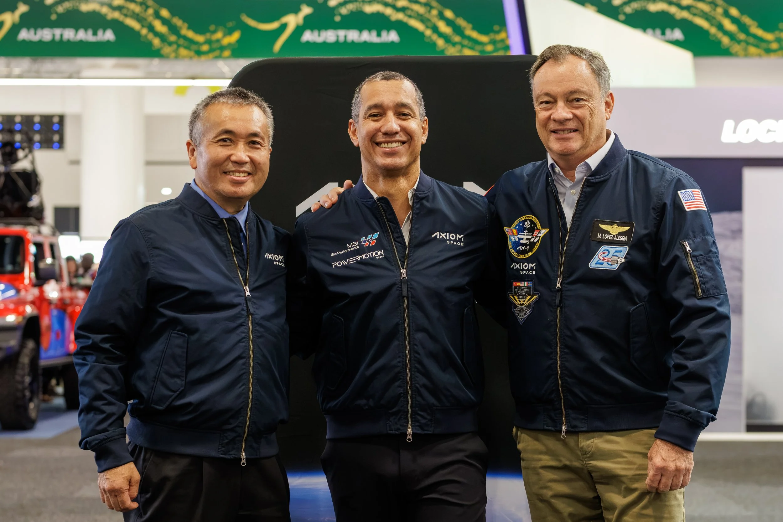Three men dressed in blue jackets smiling, standing together at an indoor event with Australian flags and space-themed patches on their jackets.