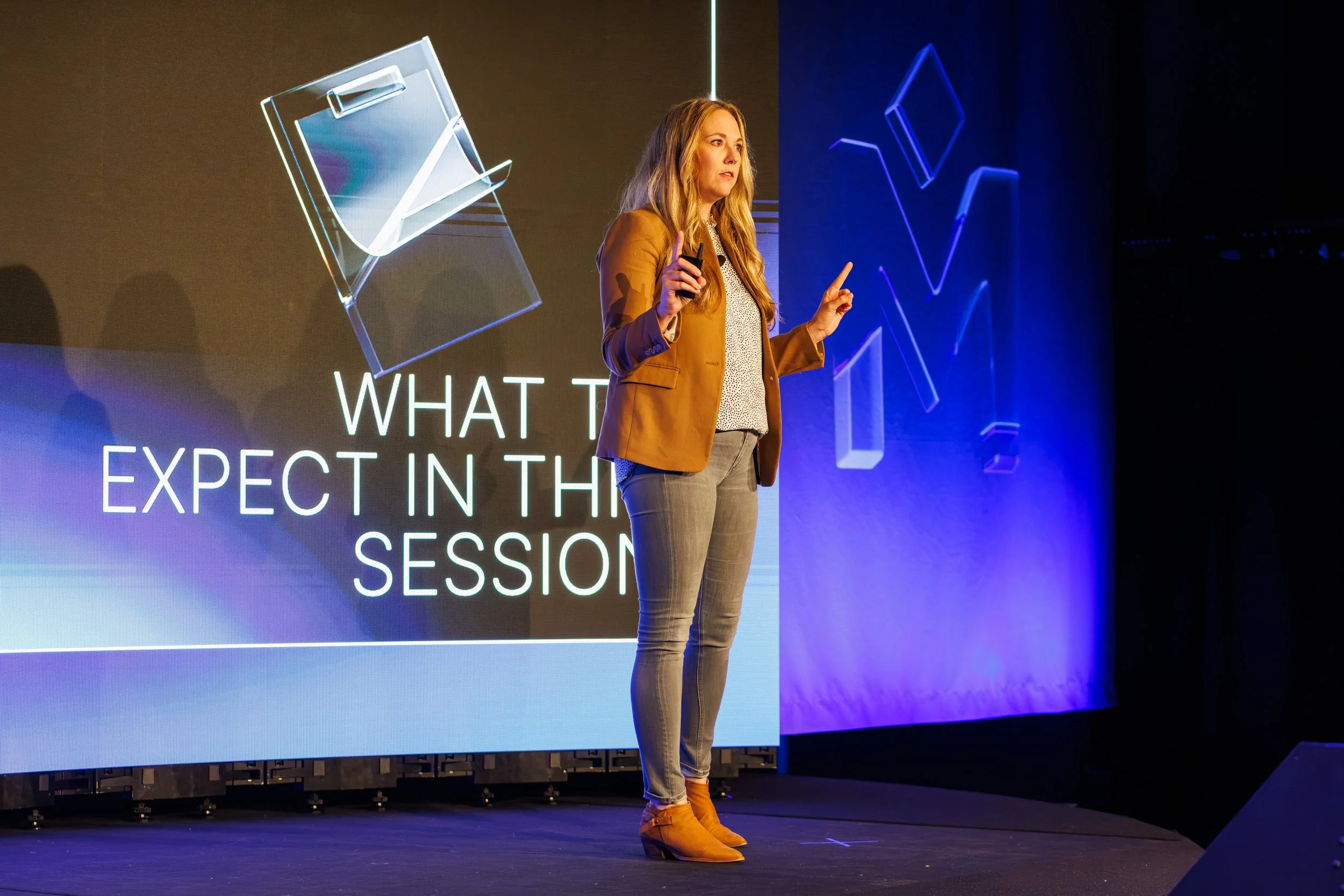 A woman giving a presentation on stage with a large screen behind her displaying the words 'What to expect in the session' and abstract graphic designs.