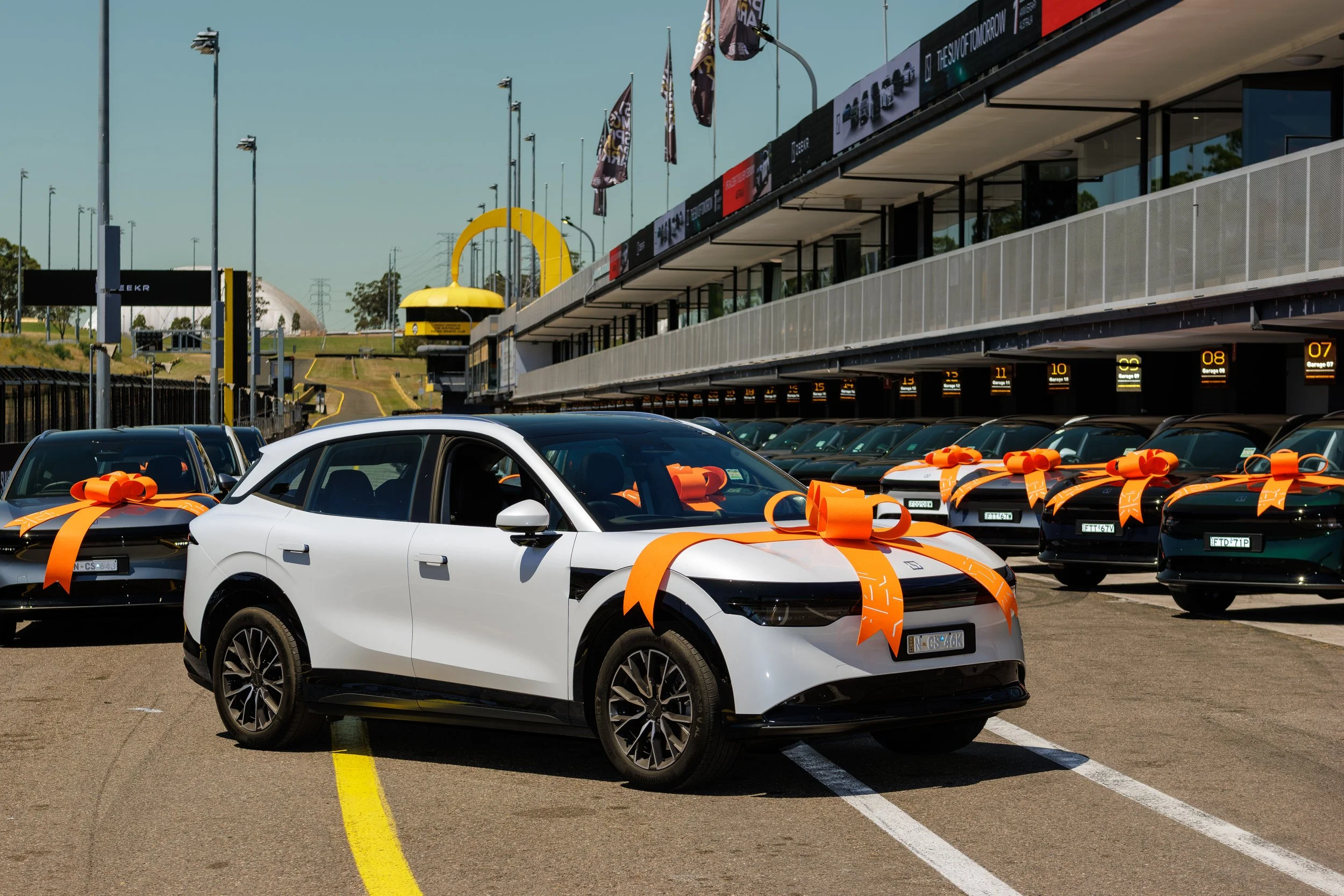 Multiple cars with orange ribbons parked at a race track, with a large yellow arch, flags, and digital signage in the background.
