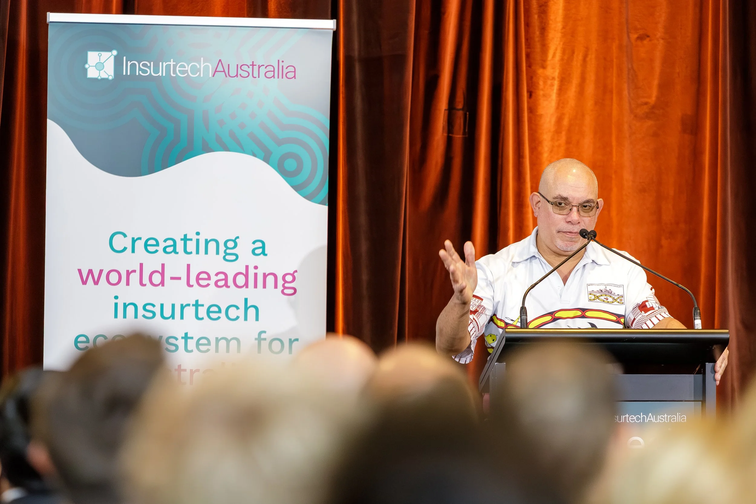 A man speaking at a podium during a conference with a banner beside him that reads 'Insurtech Australia' and 'Creating a world-leading insurtech ecosystem for'.