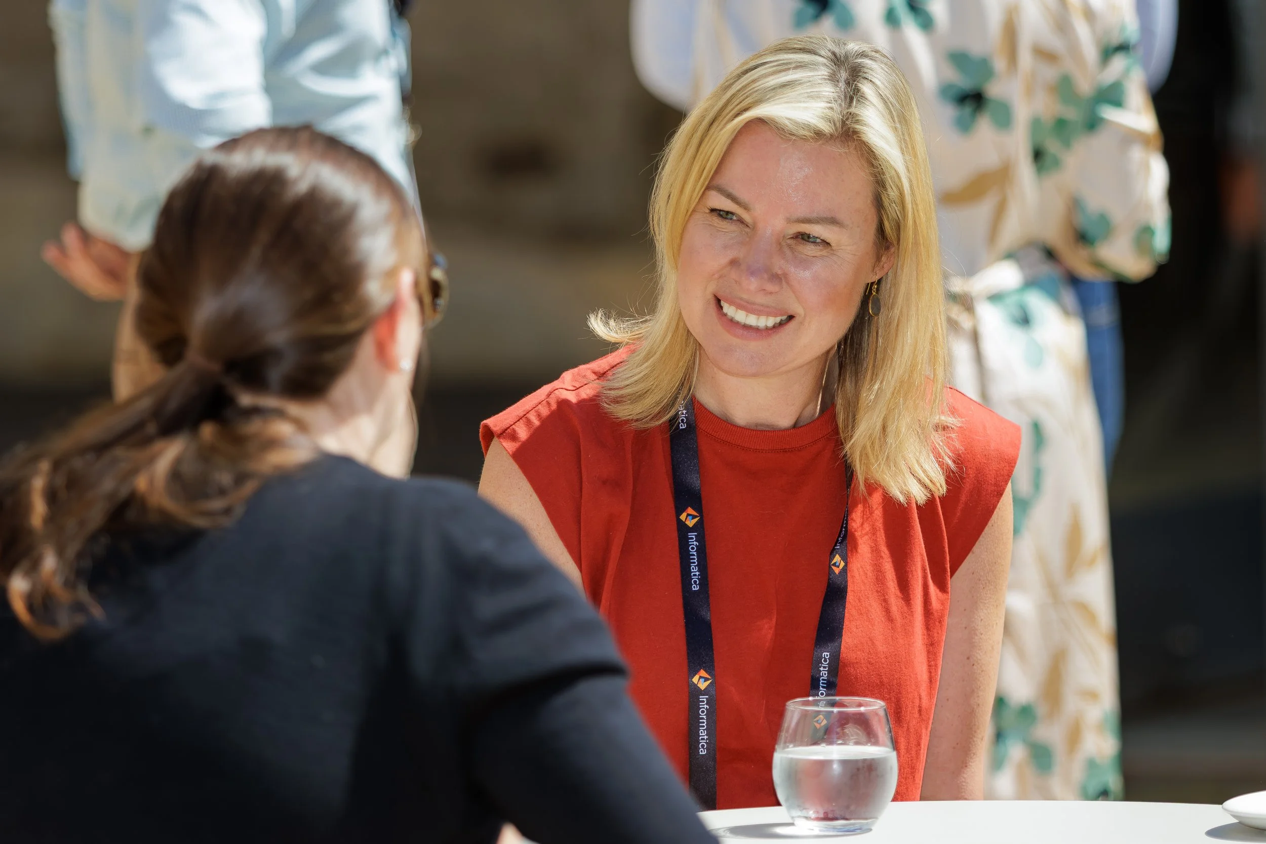 A blonde woman in a red shirt is engaged in conversation with another woman with brown hair at a social gathering, with a glass of water on the table in front of her.