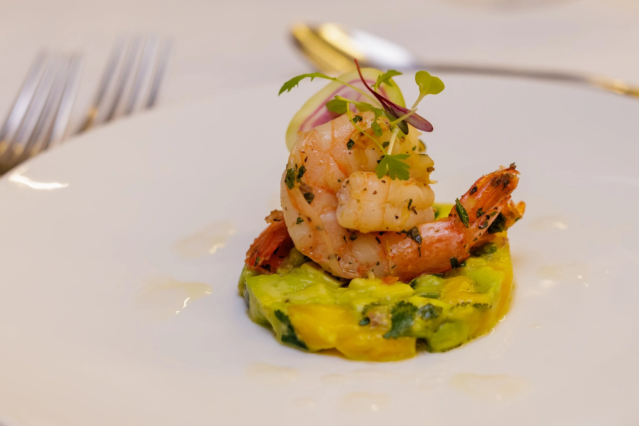 Close-up of a plated seafood dish with shrimp and garnish on a bed of sliced vegetables, with cutlery in the background.