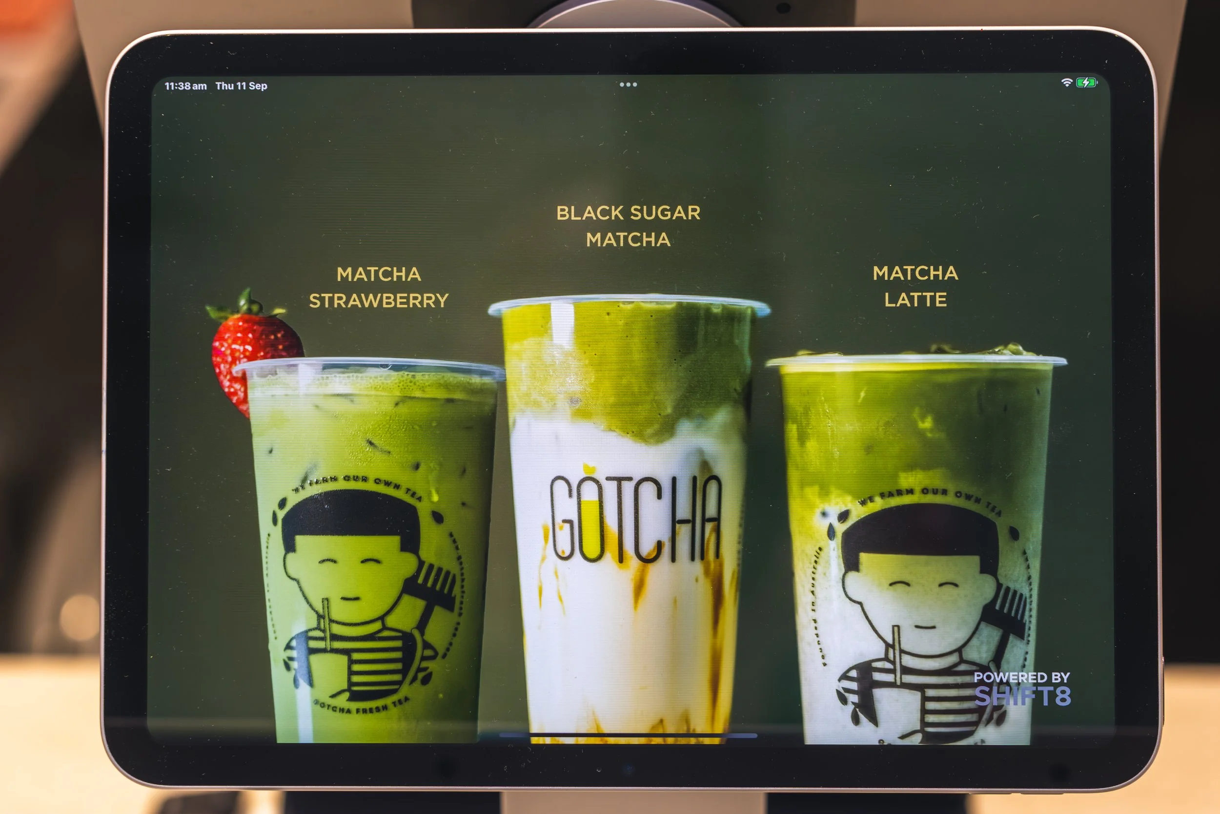 Three green matcha drinks in clear plastic cups with black and yellow branding, one topped with a strawberry, on a digital screen. The screen displays a menu with options such as Black Sugar Matcha, Matcha Strawberry, and Matcha Latte. The date and t