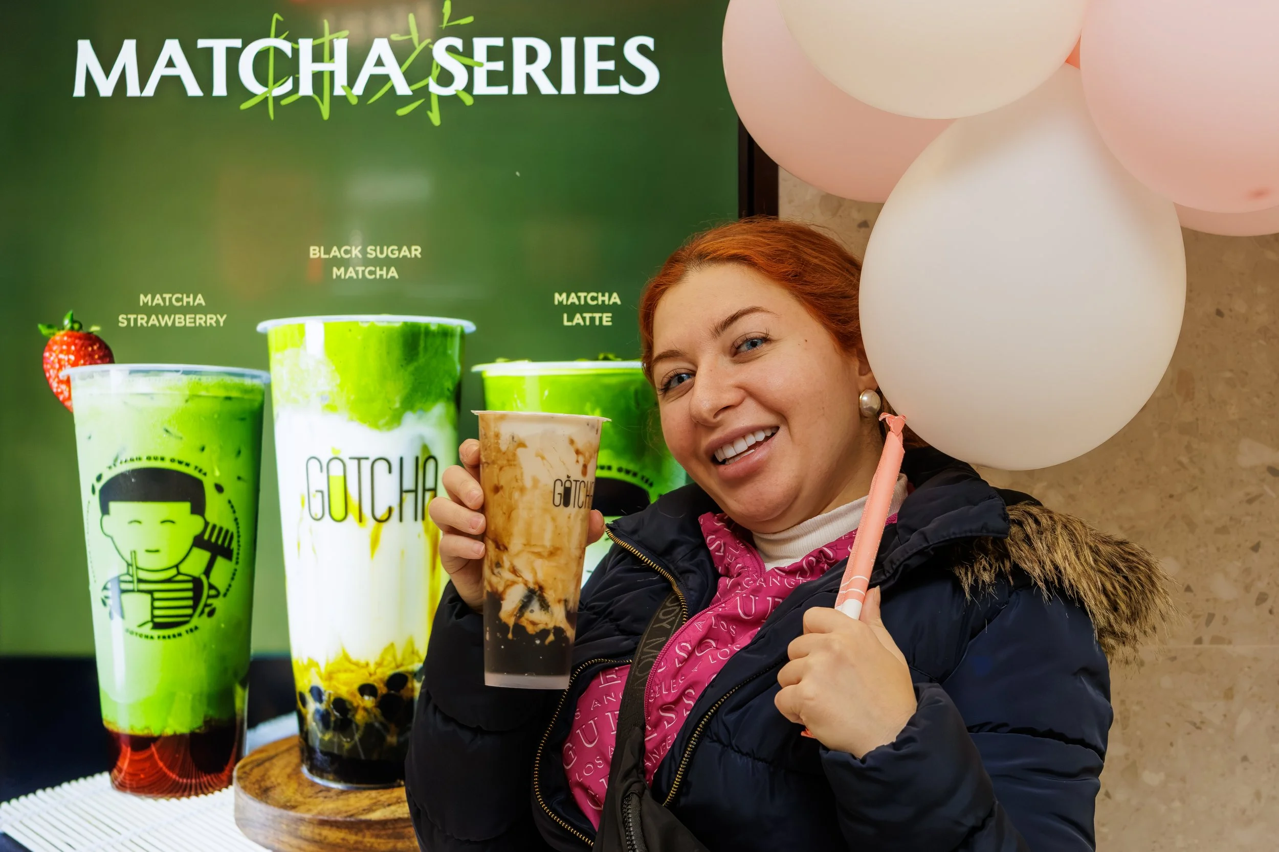 A woman with red hair and earrings smiling and holding a bubble tea drink with brown swirls and tapioca pearls, standing in front of a promotional display for Matcha Series beverages that includes large images of cups of matcha drinks and balloons.