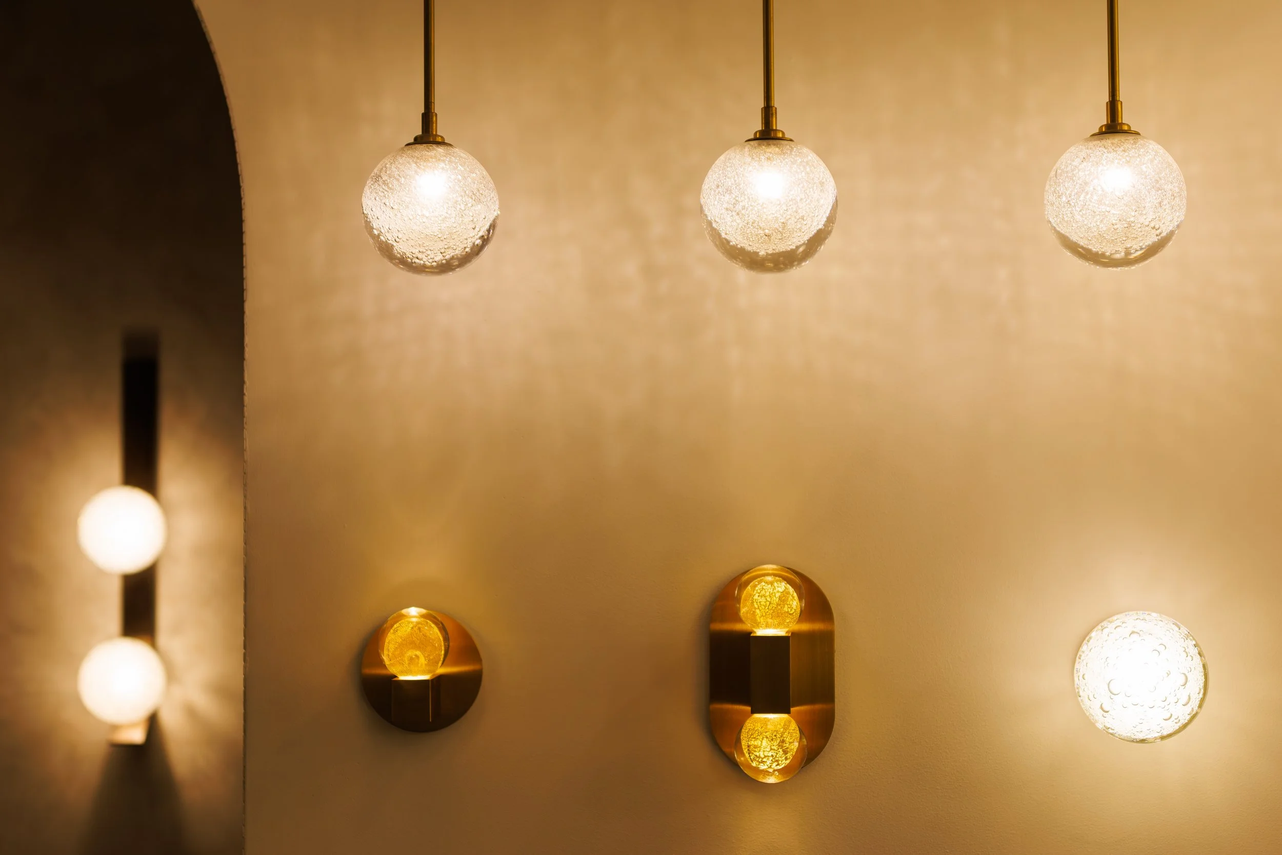 A series of modern wall-mounted sconce lights with spherical glass shades and gold accents, mounted on a beige wall.