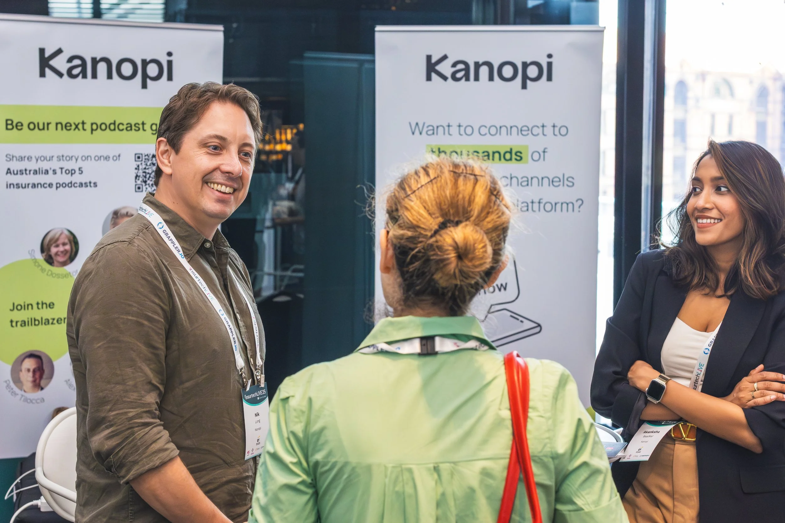 A group of four people talking at a professional event or conference, with promotional banners and badges visible.