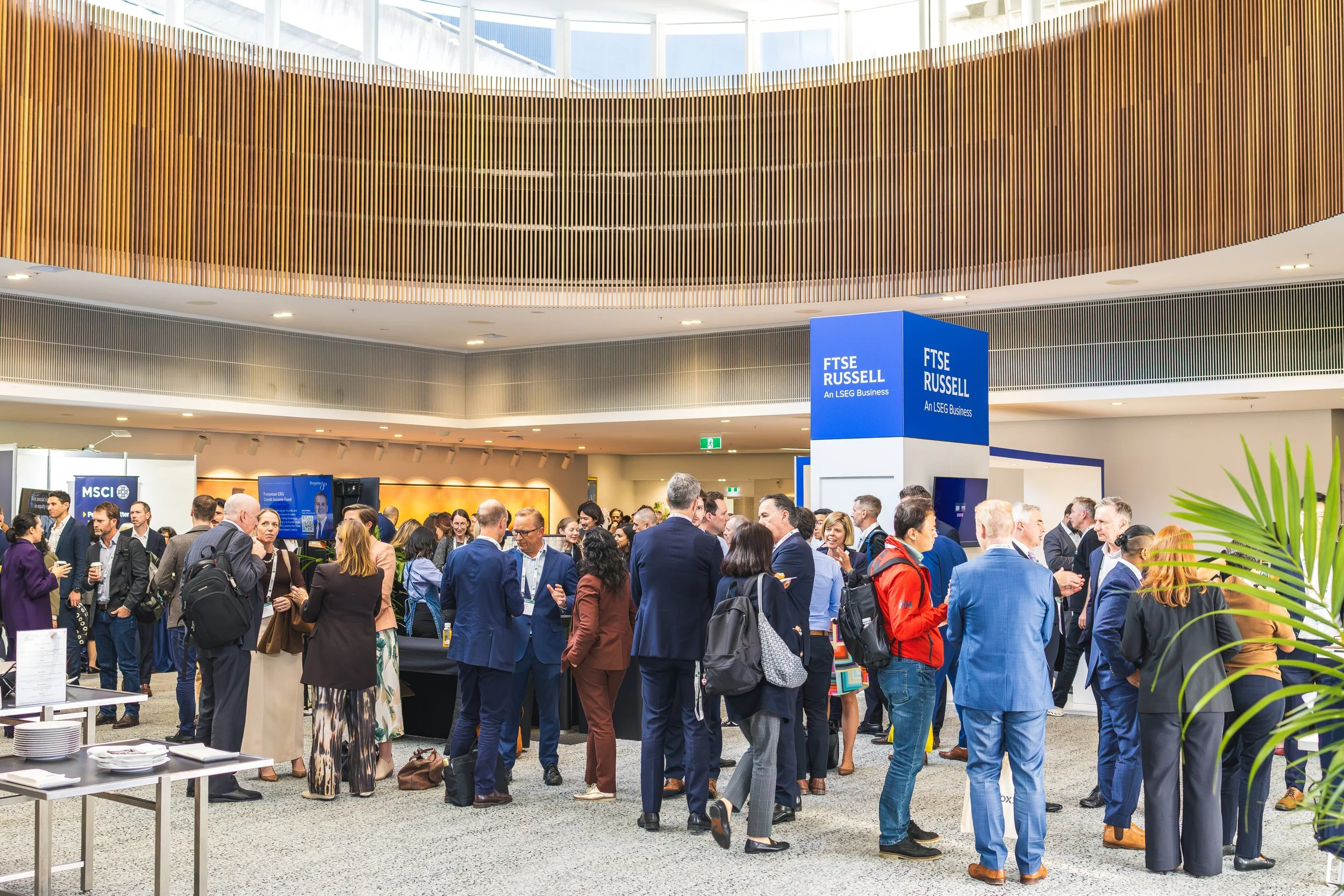A crowded professional networking event or conference inside a modern, spacious building with wooden architectural accents. Attendees are interacting and gathering near display booths labeled 'FTSE RUSSELL' and 'MSCI', with some holding drinks and en