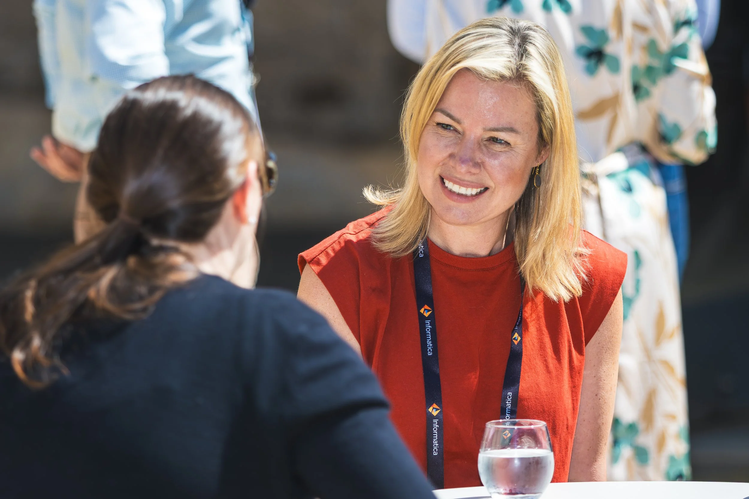 A woman with blonde hair smiling while talking to another woman with brown hair at a social event, with a glass of wine on the table in front of her.