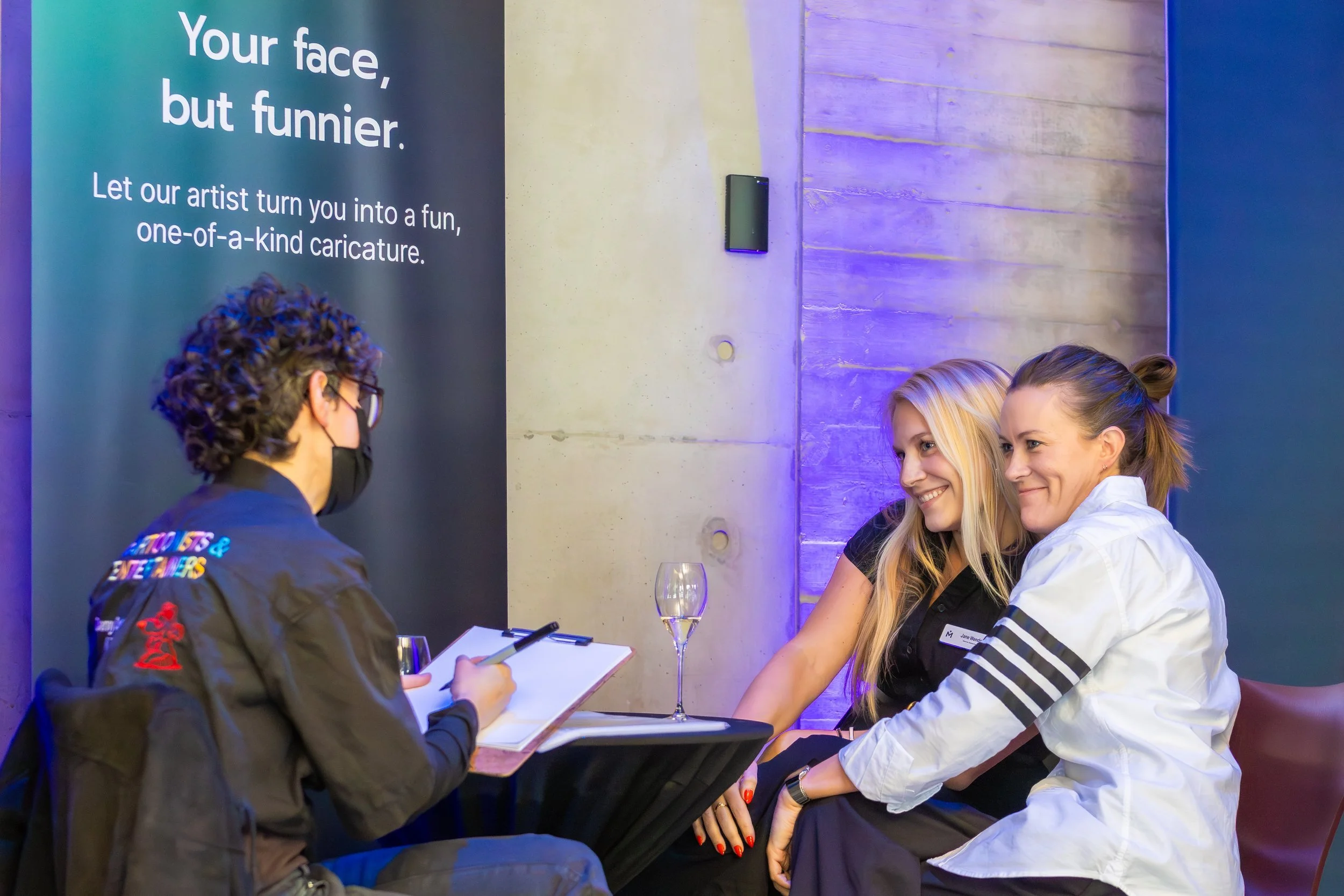 People sitting at a table during a caricature drawing event, with a sign that says, 'Your face, but funnier. Let our artist turn you into a fun, one-of-a-kind caricature.'