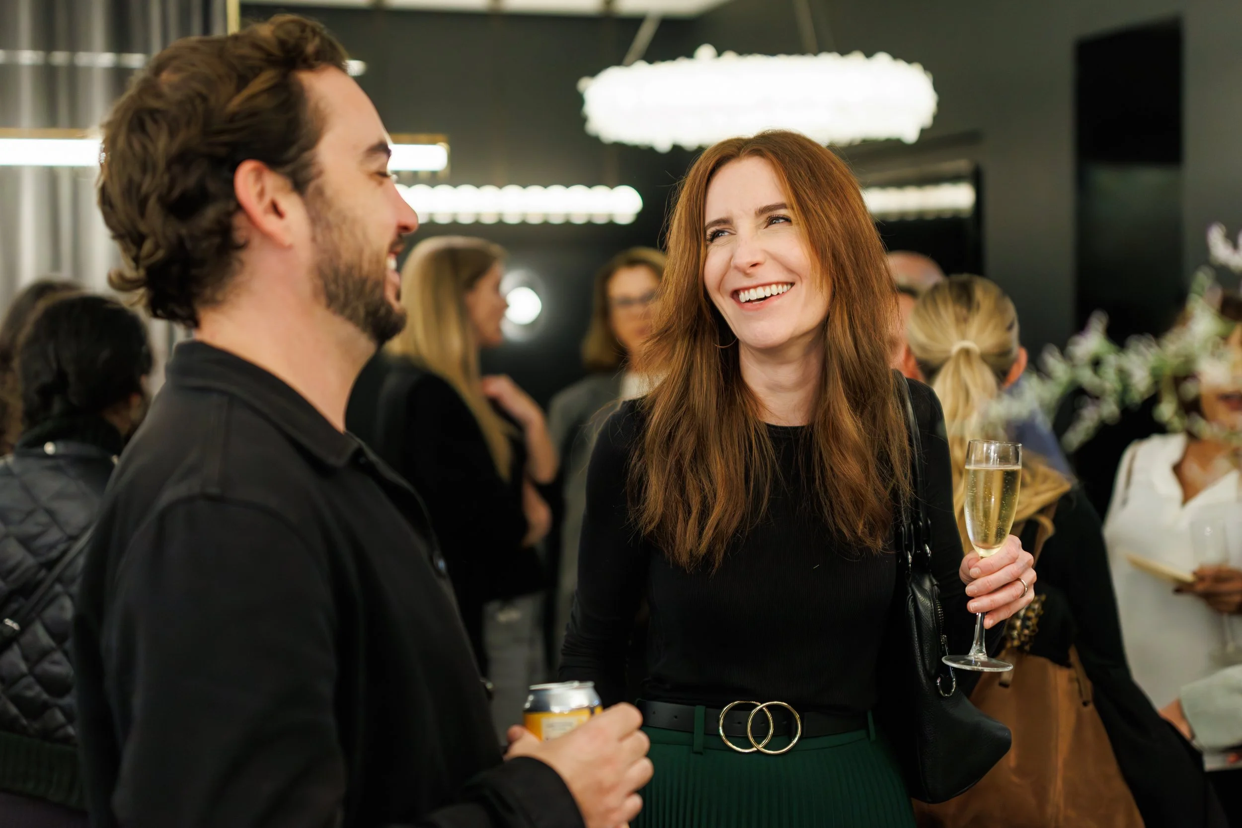 A woman with red hair smiling while holding a glass of champagne at a social gathering, talking to a man with dark hair and a beard.