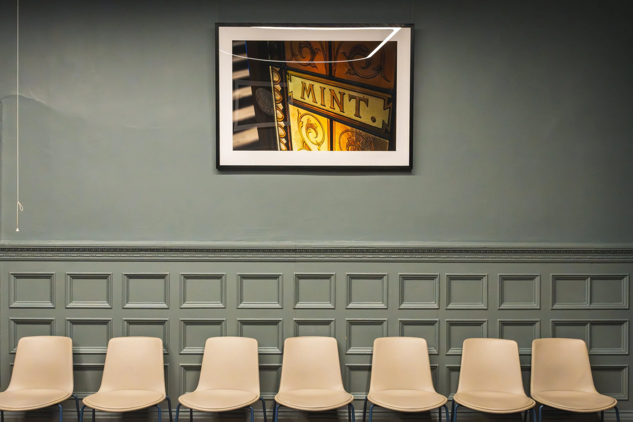 Empty beige chairs in front of a gray wall with decorative paneling, and a framed stained glass window with the word 'MINT' visible.
