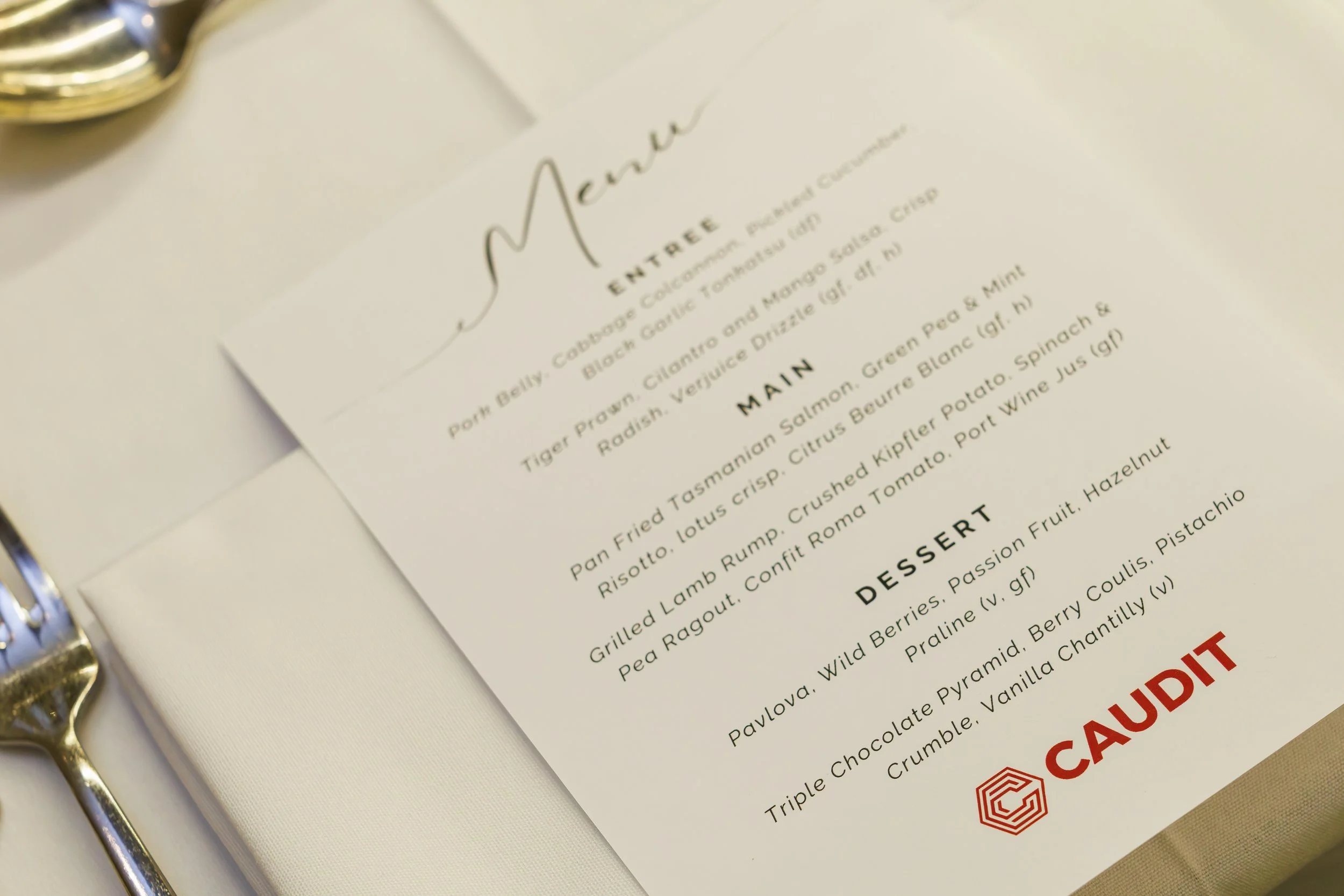 Close-up of a restaurant menu on a table, with visible sections titled 'ENTREE', 'MAIN', and 'DESSERT'. The menu features dishes like tropical vegetables, grilled lamb, and assorted desserts. Part of a fork and spoon are seen on the left side of the 