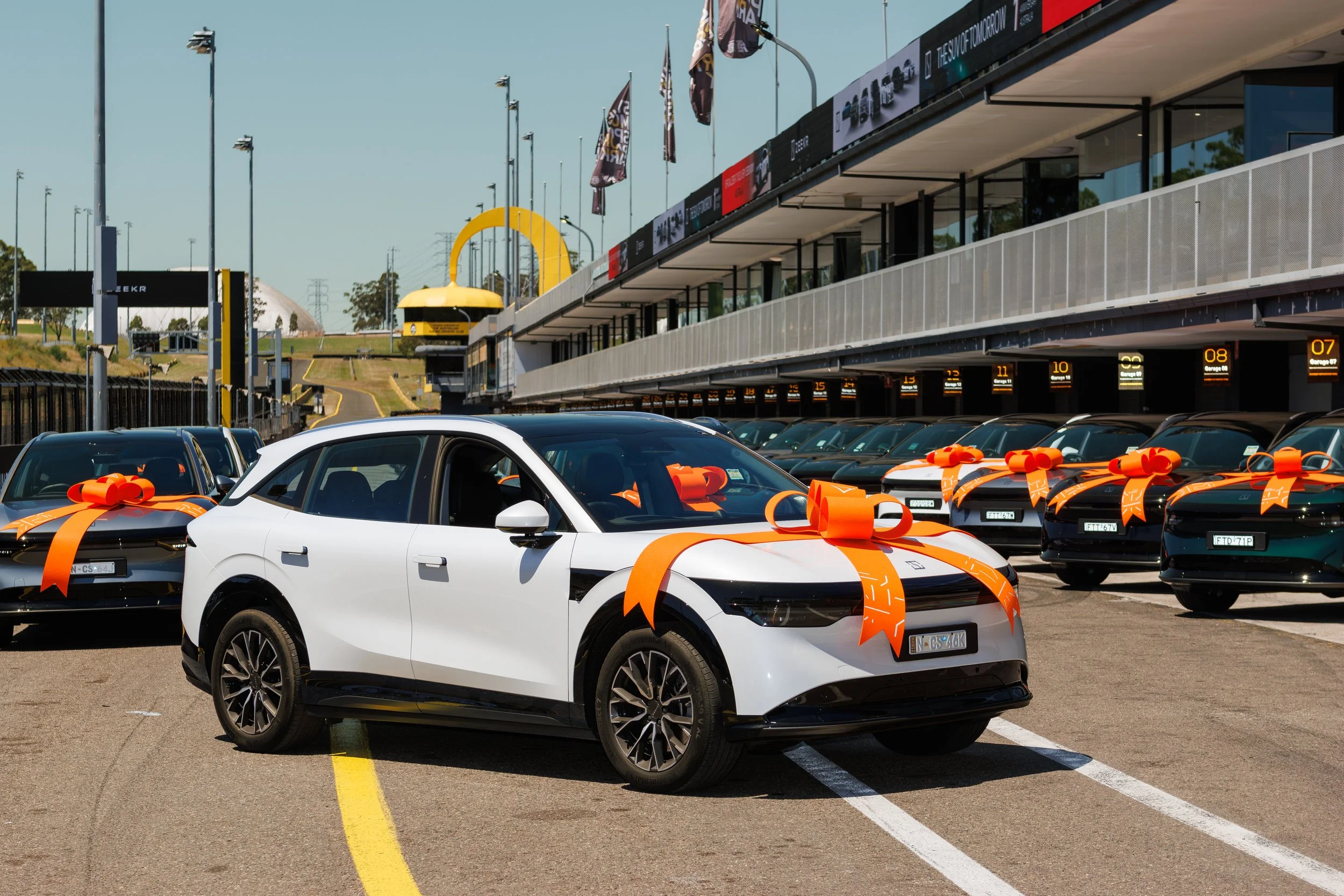 Multiple cars parked at a racetrack with orange bows on the hoods. A building with digital panels and flags is in the background, along with a large yellow sculpture and a clear sky.