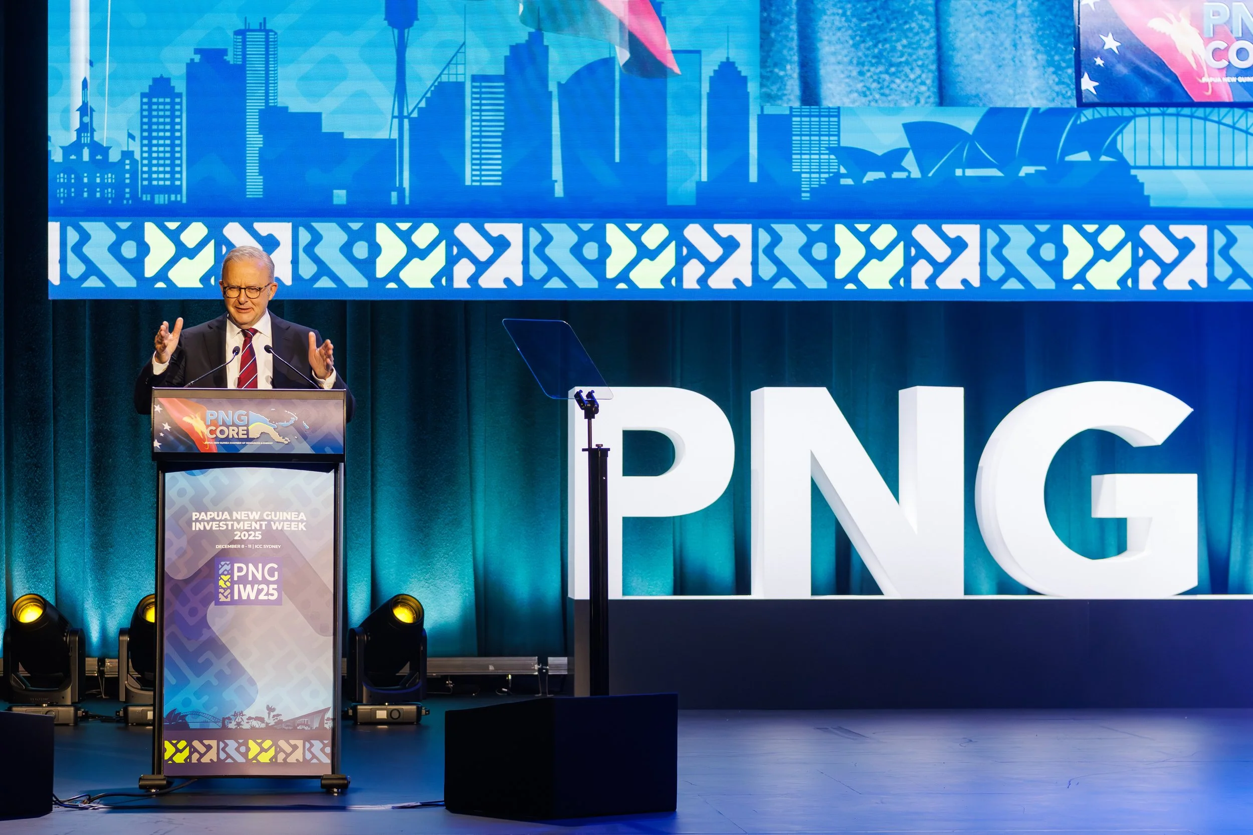 A man with glasses and gray hair is speaking at a podium. There are large illuminated letters behind him spelling 'PNG.' The background features a large blue cityscape graphic and part of a sign for Papua New Guinea Investment Week 2025.