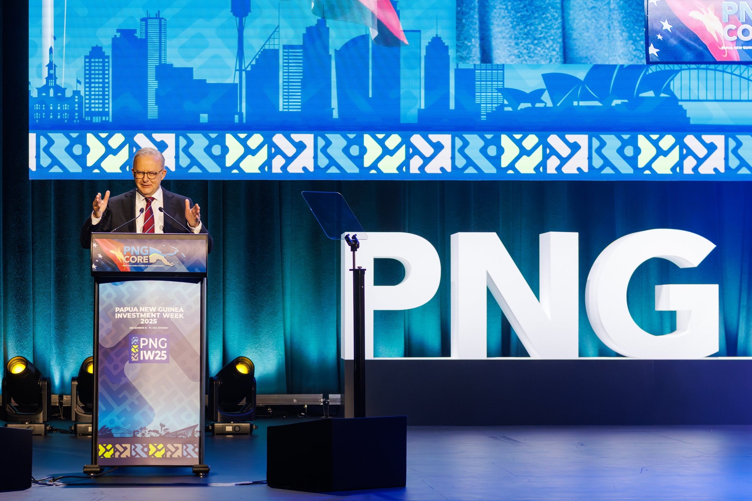 A man with glasses and gray hair is speaking at a podium. There are large illuminated letters behind him spelling 'PNG.' The background features a large blue cityscape graphic and part of a sign for Papua New Guinea Investment Week 2025.