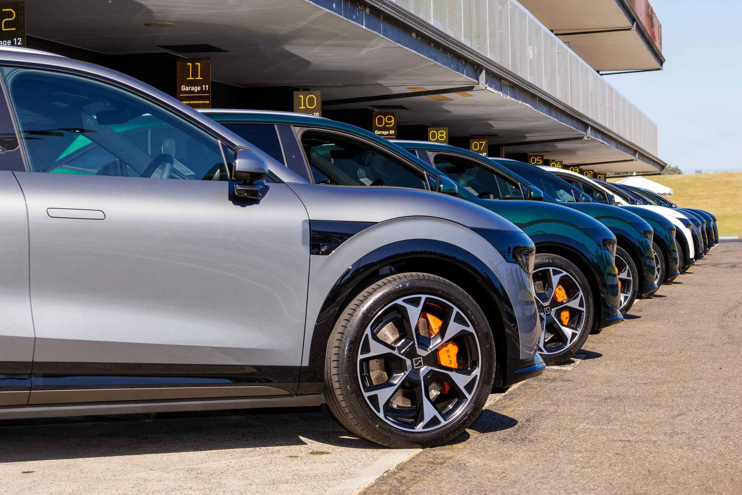 Multiple cars parked in a row at a racing circuit's garage area, with garage numbers visible above each car.