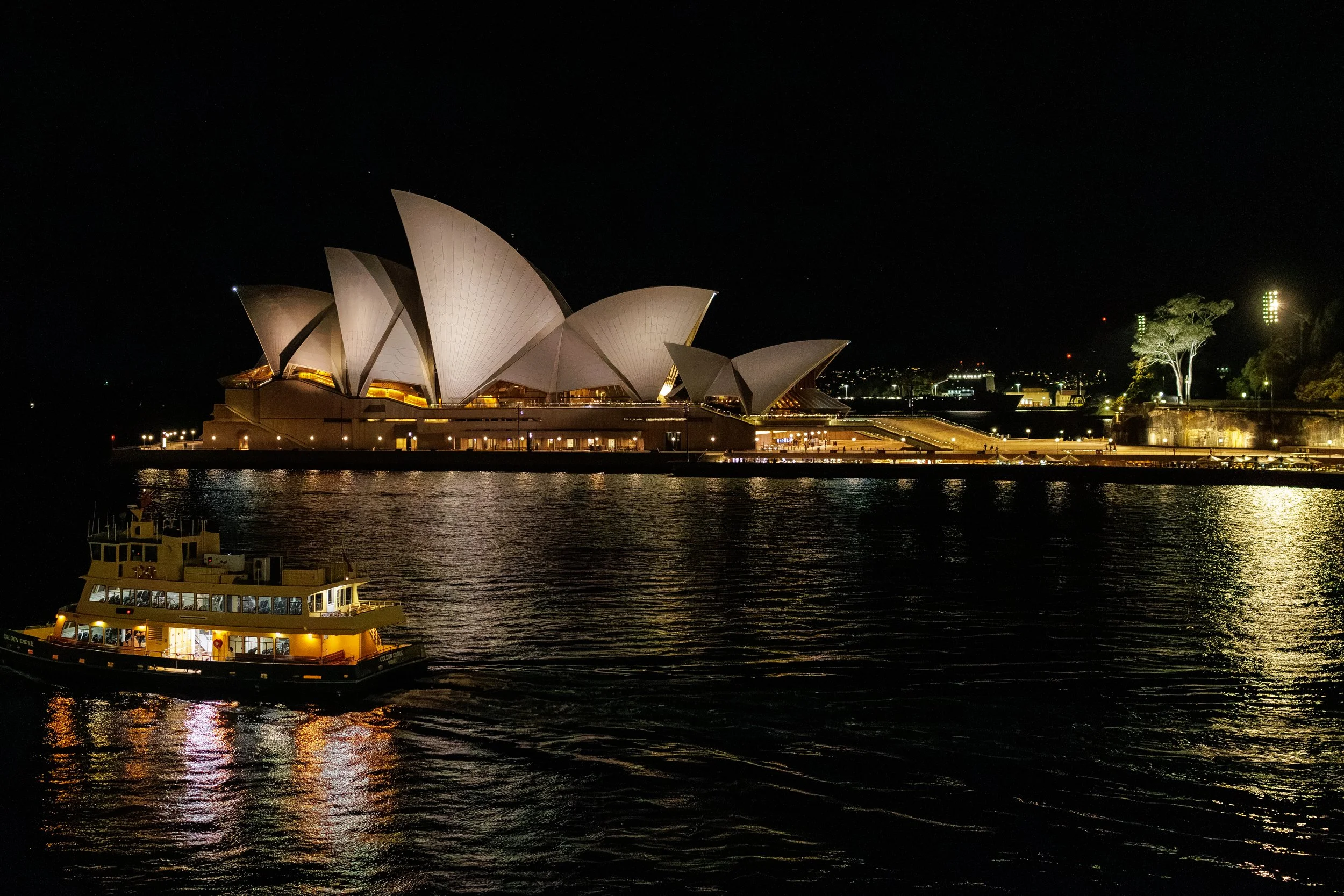 Nighttime view of the Sydney Opera House illuminated, reflecting on the water with a ferry boat in the foreground.
