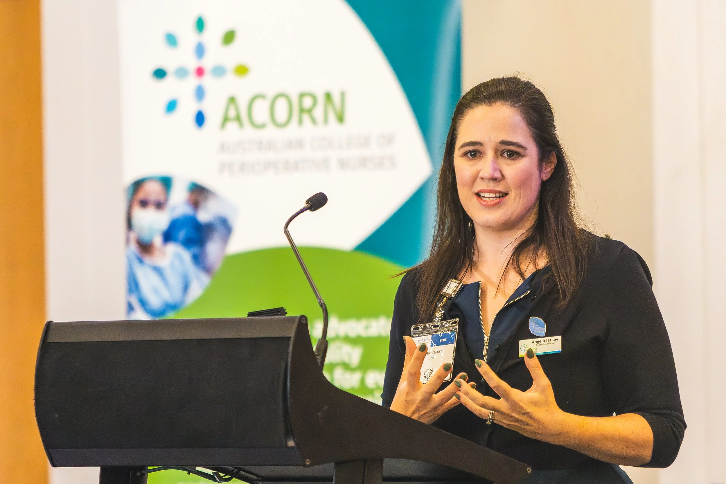 A woman with long dark hair, wearing a black jacket and name tag, is speaking at a podium with a microphone in front of a colorful banner with the text 'ACORN Australian College of Perioperative Nurses.'