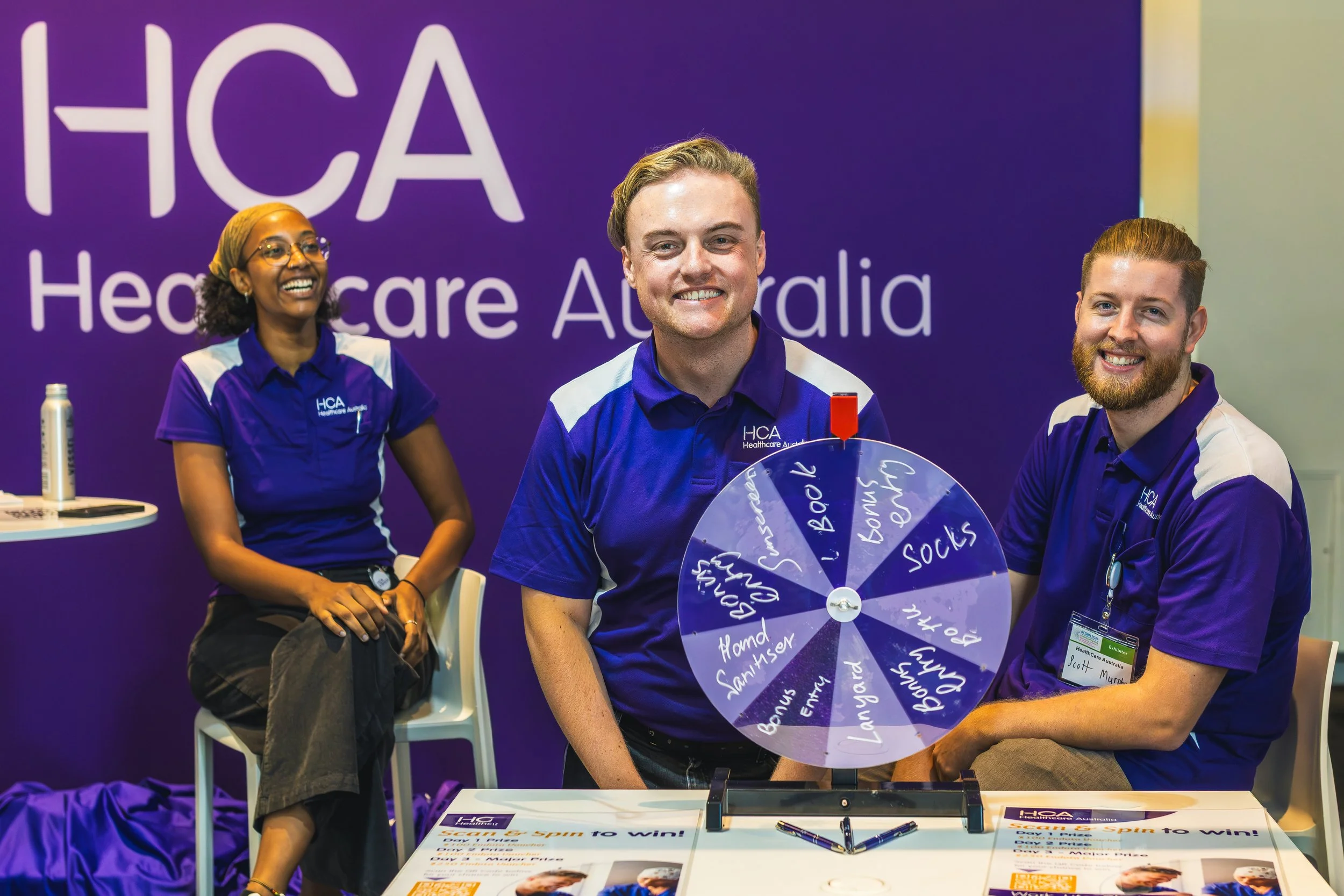 Three people sitting at a table in front of a purple HCA Healthcare Australia banner, smiling. The person in the middle holds a wheel with words like 'Bonuses,' 'Entry,' and 'Socks.' The woman on the left wears glasses, a purple shirt, and a head cov