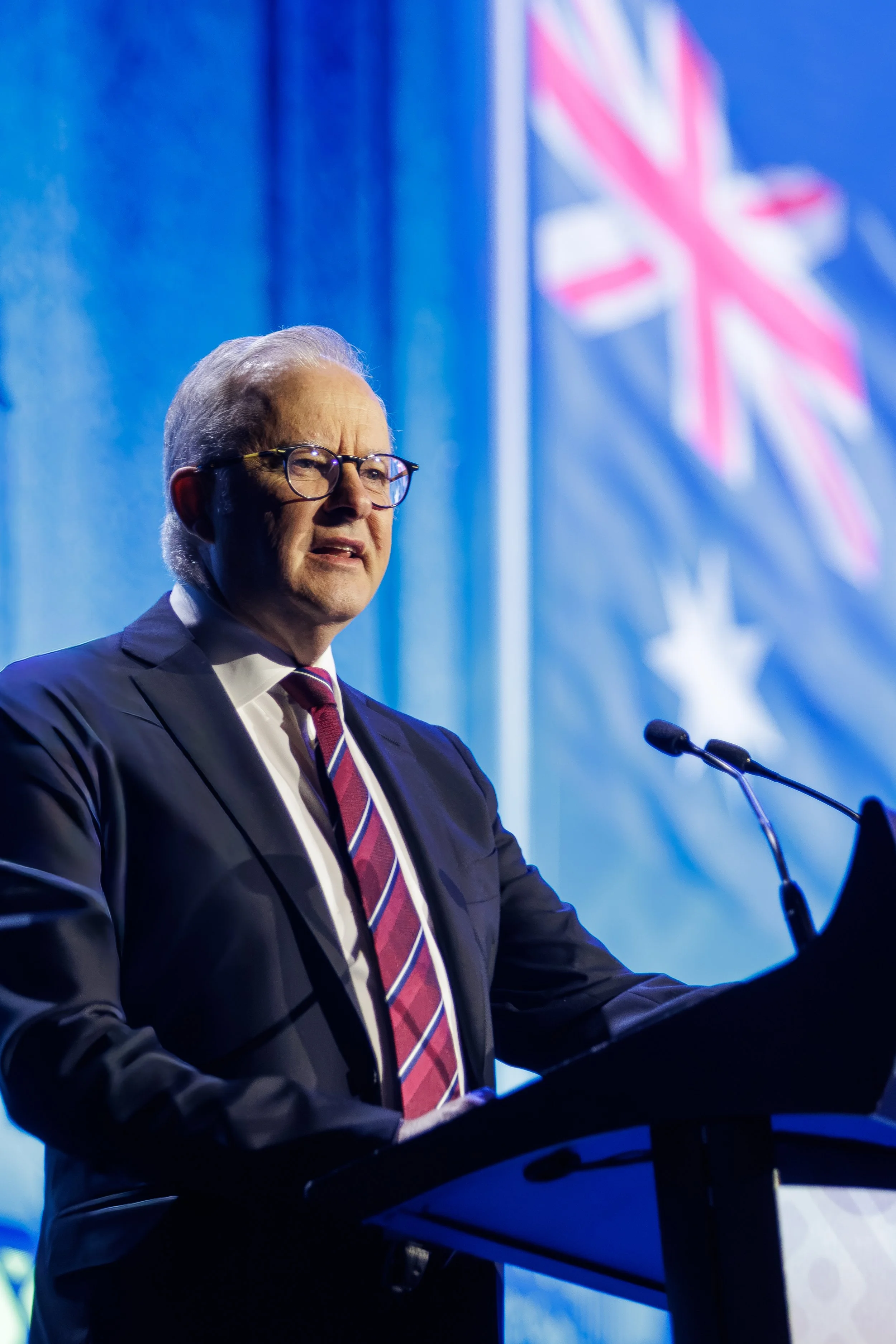 An elderly man wearing glasses and a dark suit, standing behind a podium, speaking at a formal event with a Blue and pink Union Jack flag in the background.