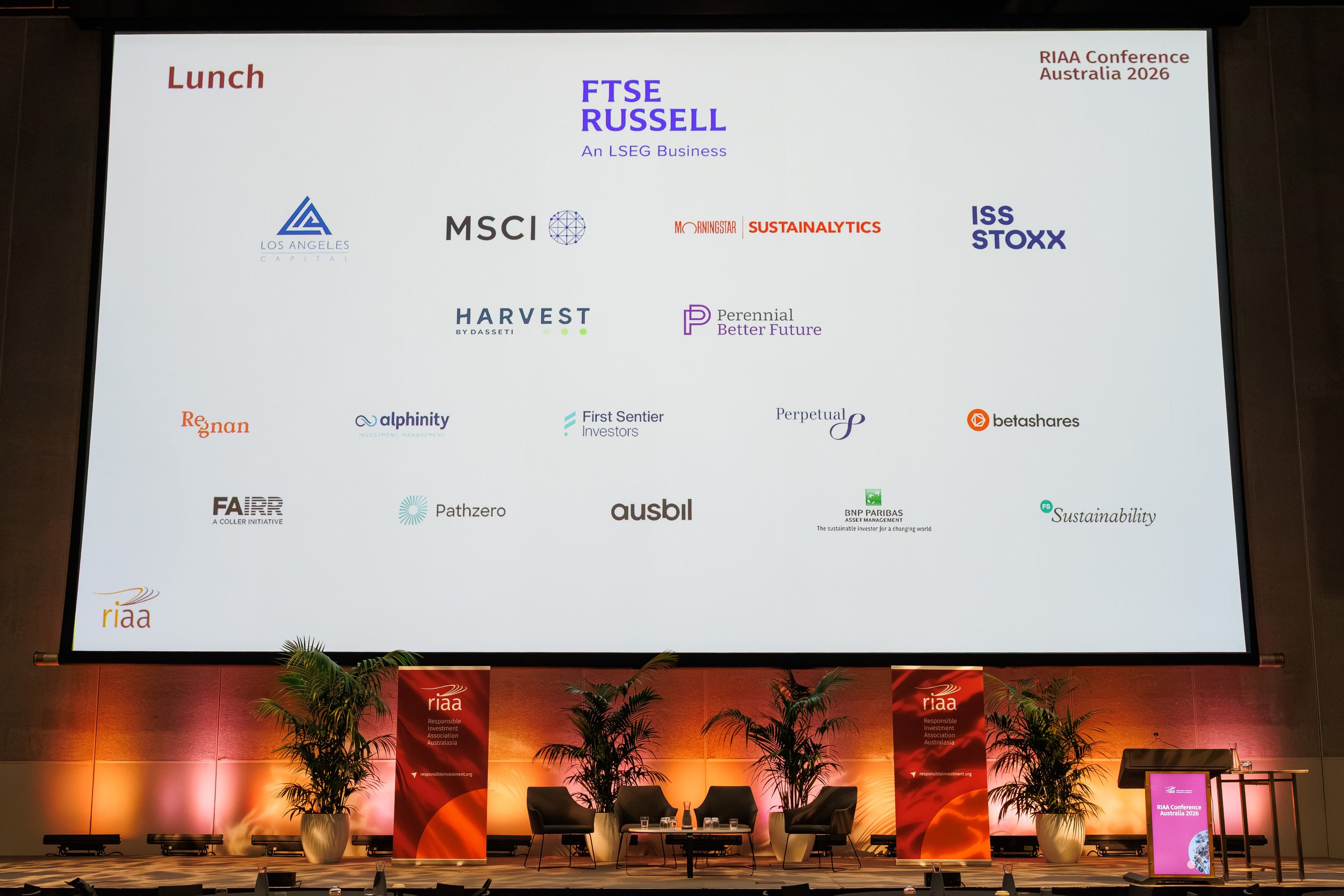 Large conference room stage set up for the RIAA Conference Australia 2026, with a big screen displaying various conference sponsors and partners, banners, plants, and chairs for speakers or panelists.