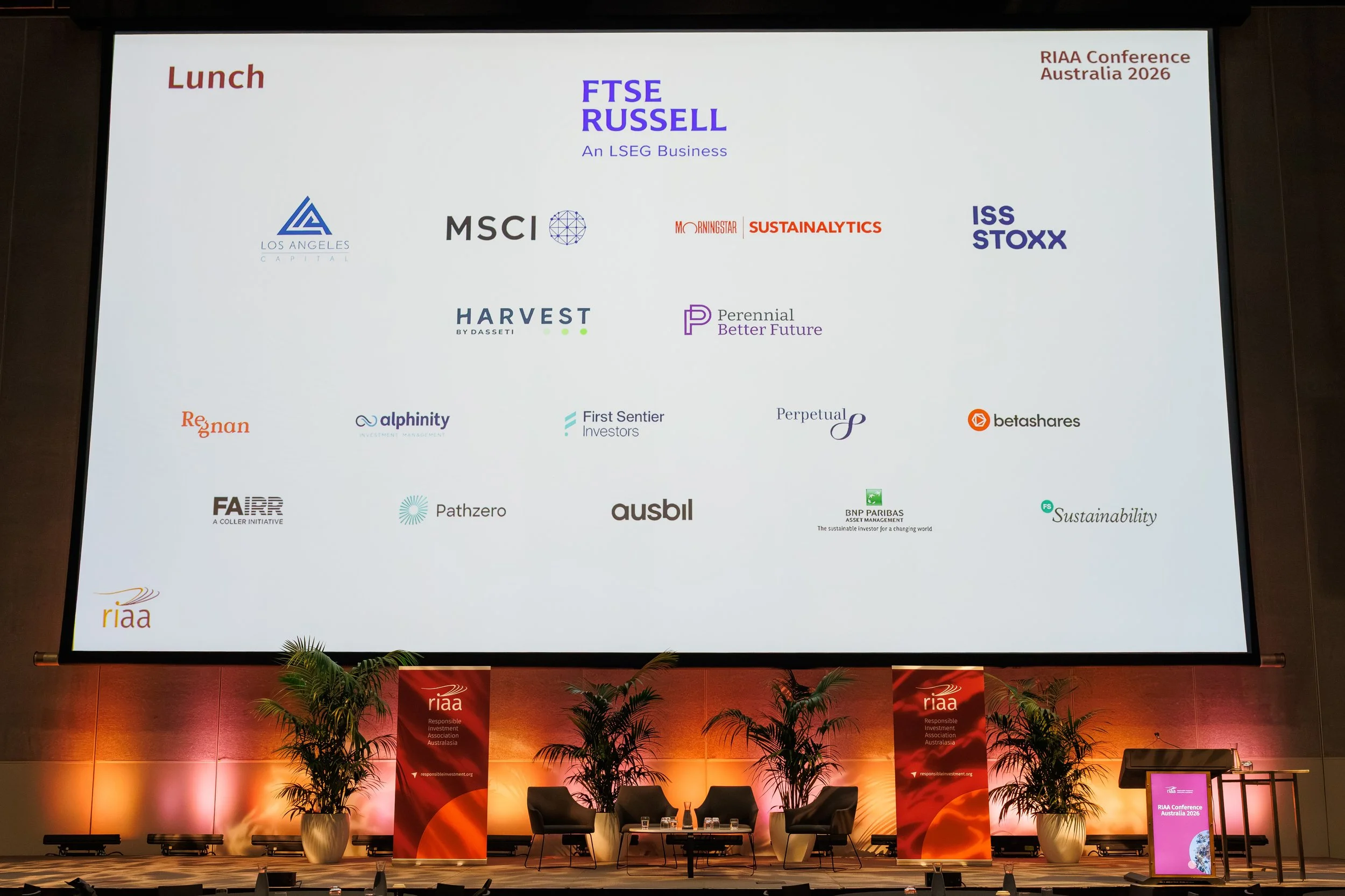 Large conference room stage set up for the RIAA Conference Australia 2026, with a big screen displaying various conference sponsors and partners, banners, plants, and chairs for speakers or panelists.