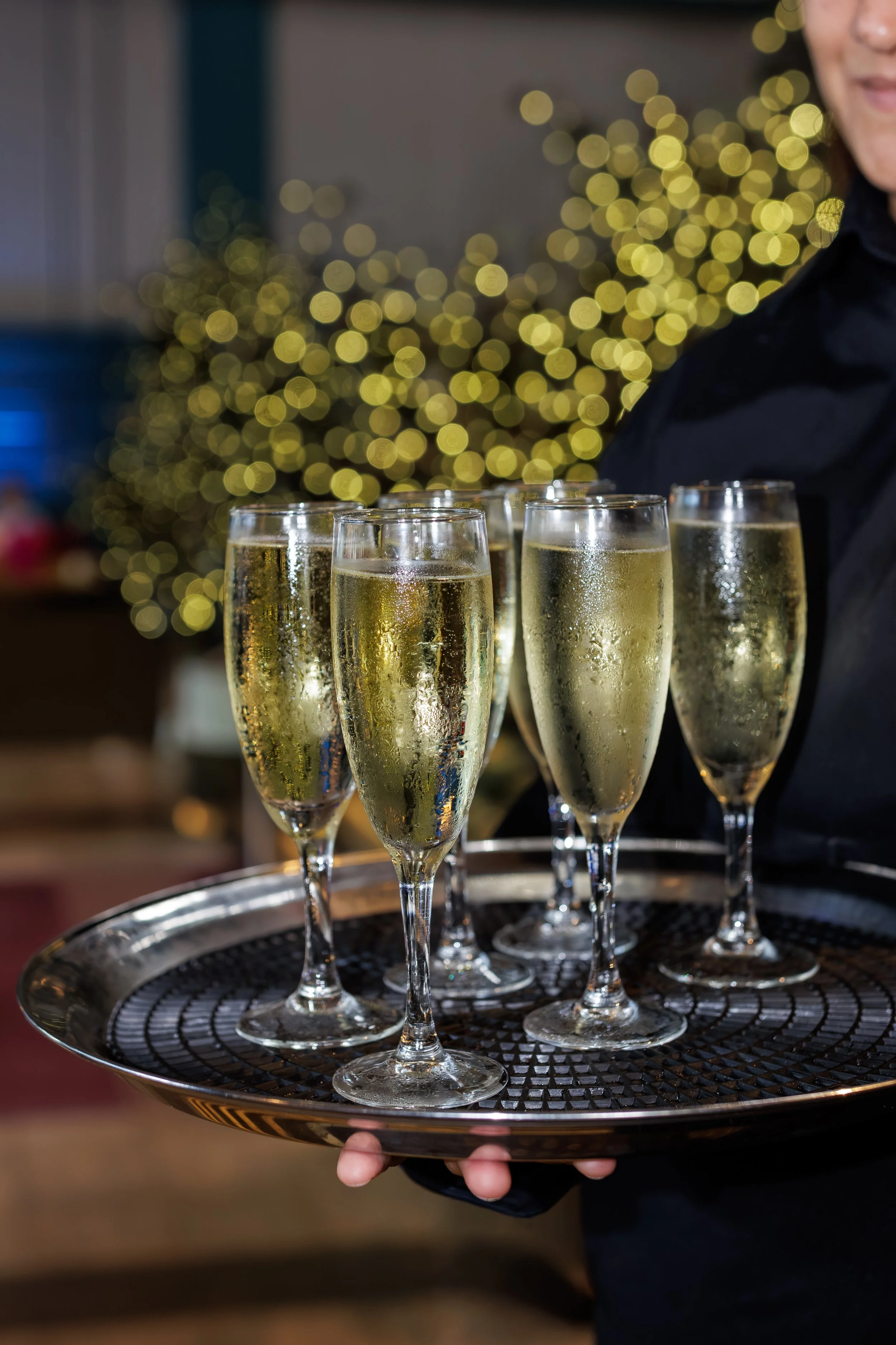 A person holding a tray of six champagne glasses filled with sparkling wine, with a decorated Christmas tree in the blurred background.