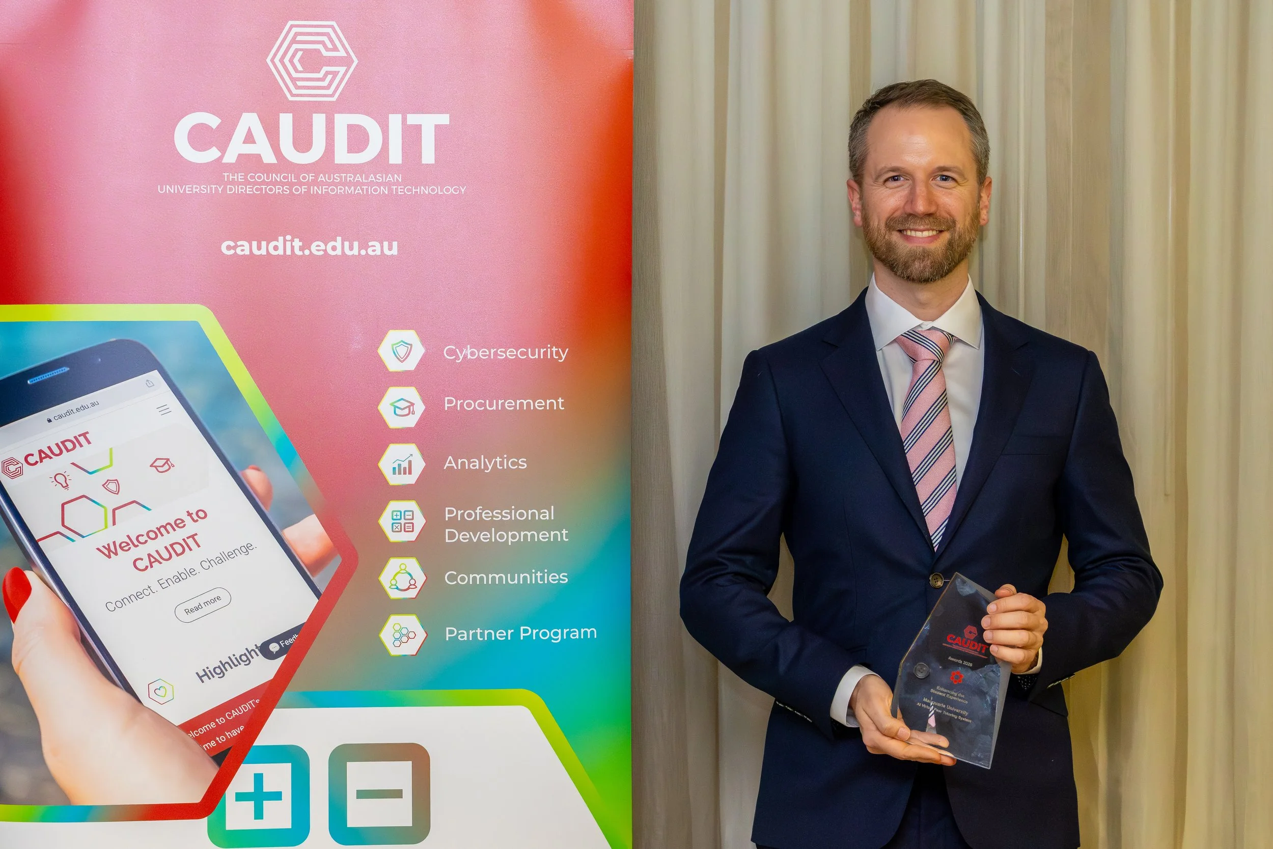 Man in a suit holding an award next to a colorful display board advertising CAUDIT, the Council of Australasian University Directors of Information Technology, with features related to cybersecurity, procurement, analytics, professional development, 