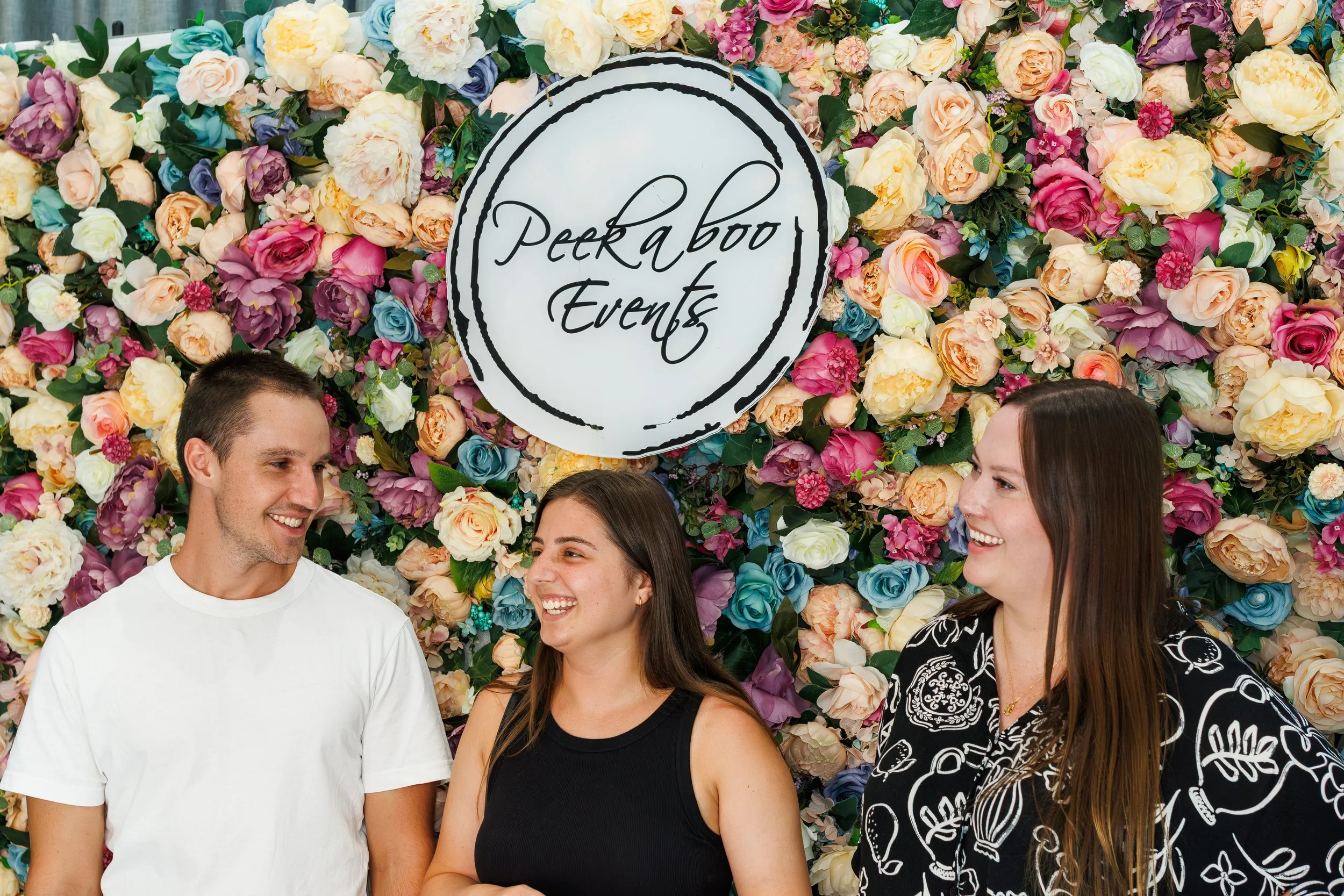 A group of four smiling people standing in front of a colorful floral wall with a sign that reads 'Peekaboo Events'.
