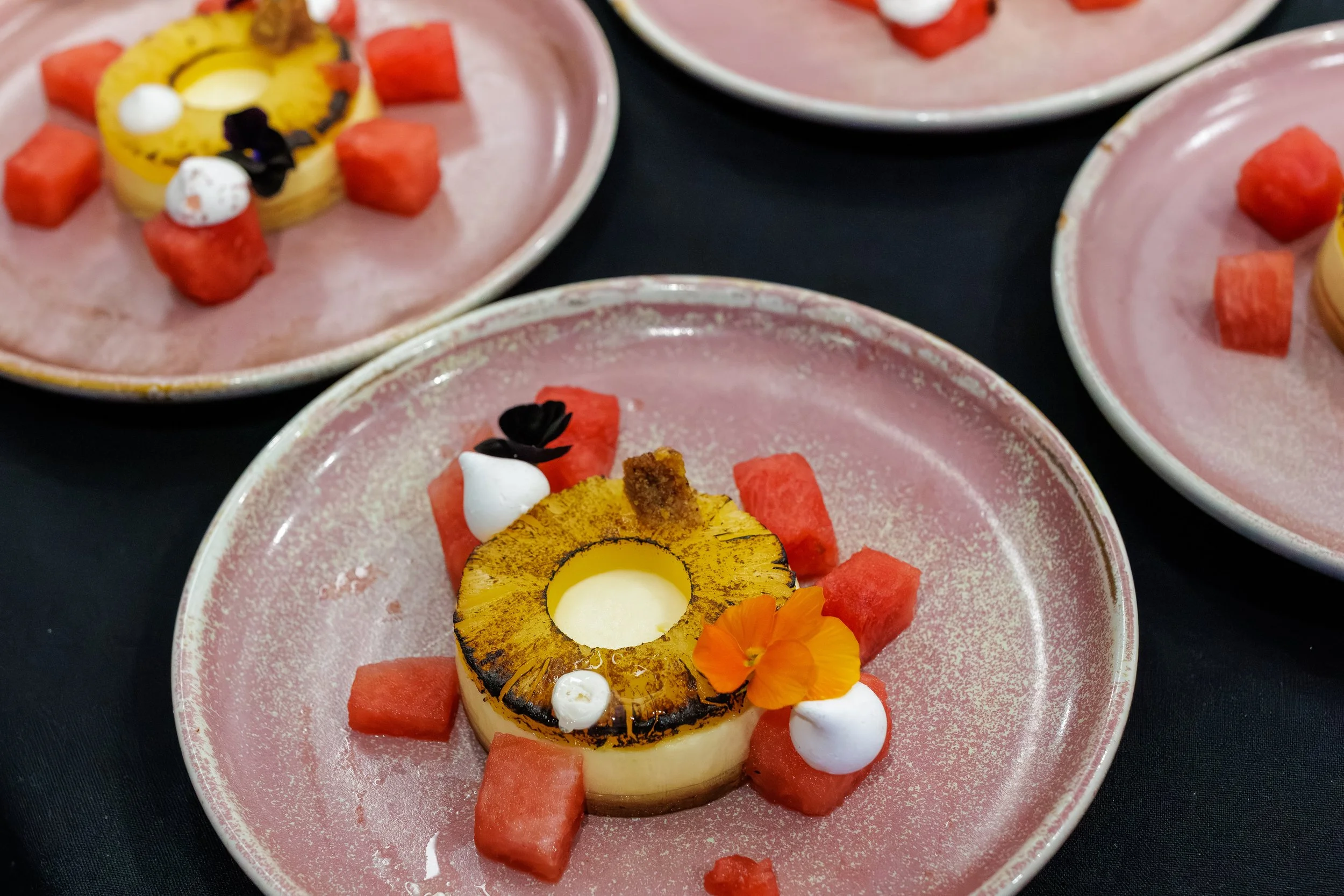 A dessert plate with grilled pineapple, watermelon cubes, and decorative dollops of white cream and small black and orange edible flowers.