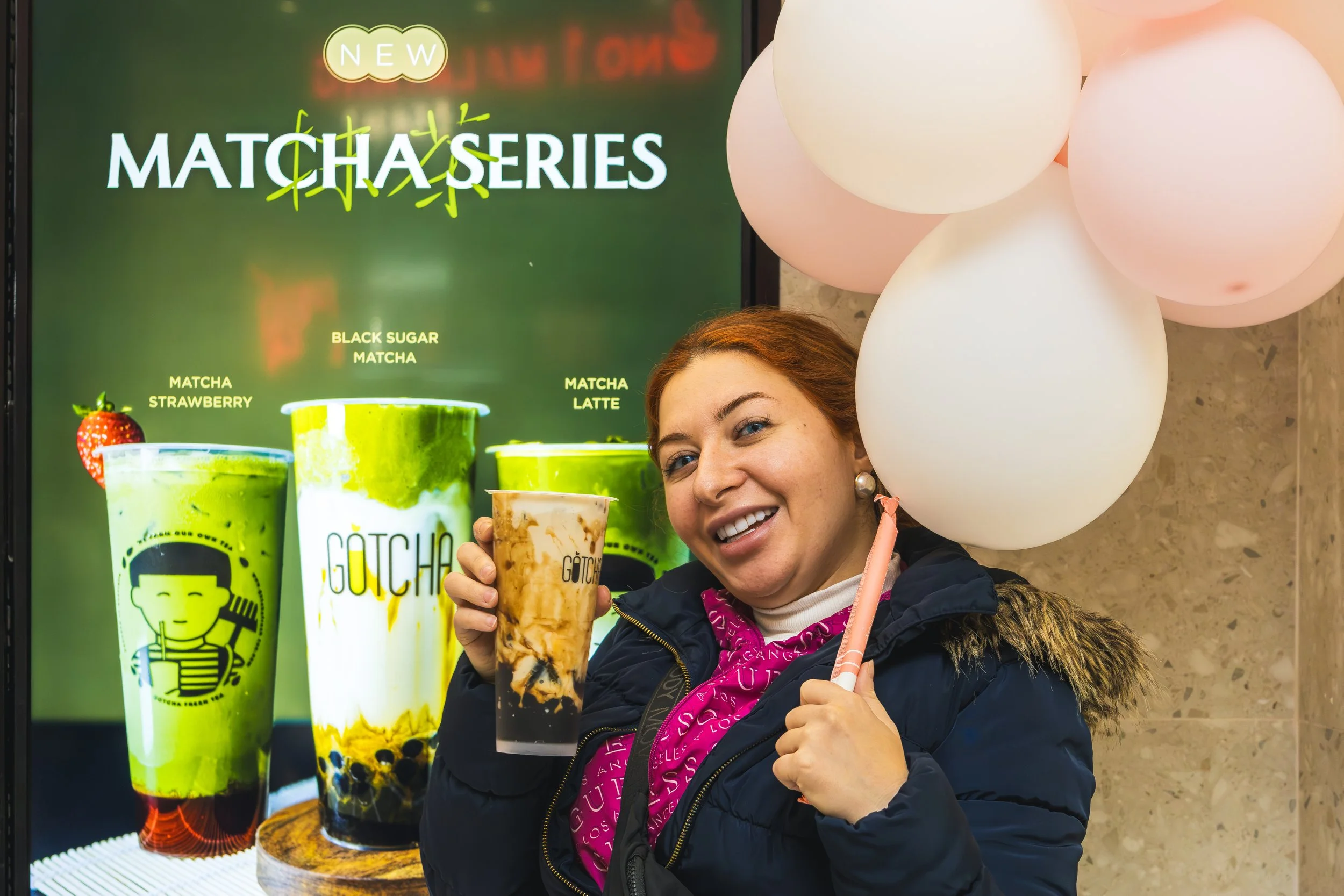A woman smiling and holding a cup of bubble tea in front of a large advertisement for Matcha Series drinks, with images of various colorful matcha beverages and balloons in the background.