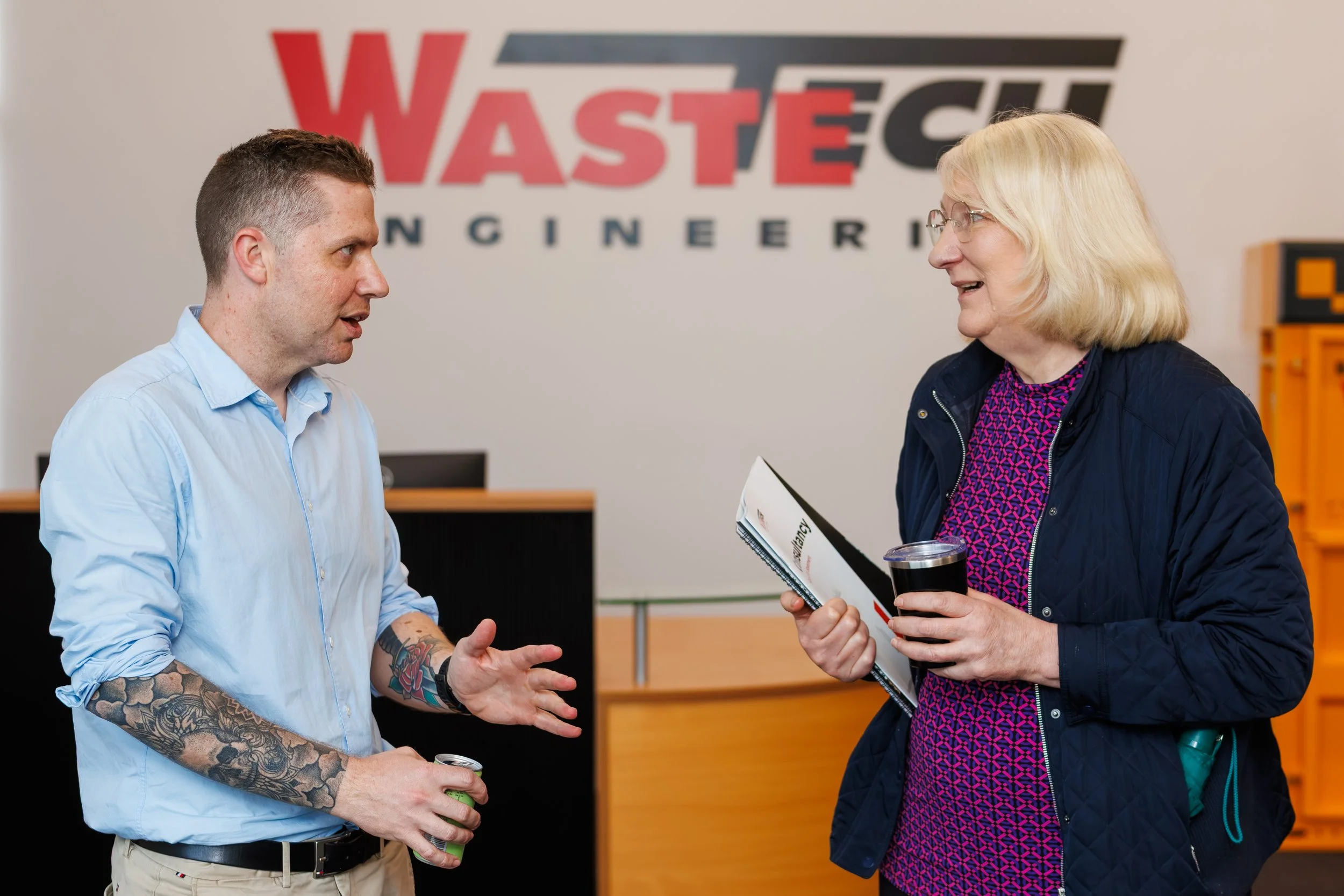 A man with tattoos on his arms and a woman with glasses having a conversation in a professional setting, standing in front of a large sign that reads 'WASTE SOLUTIONS ENGINEERING'. The woman is holding a folder and a drink, and they are engaged in a 