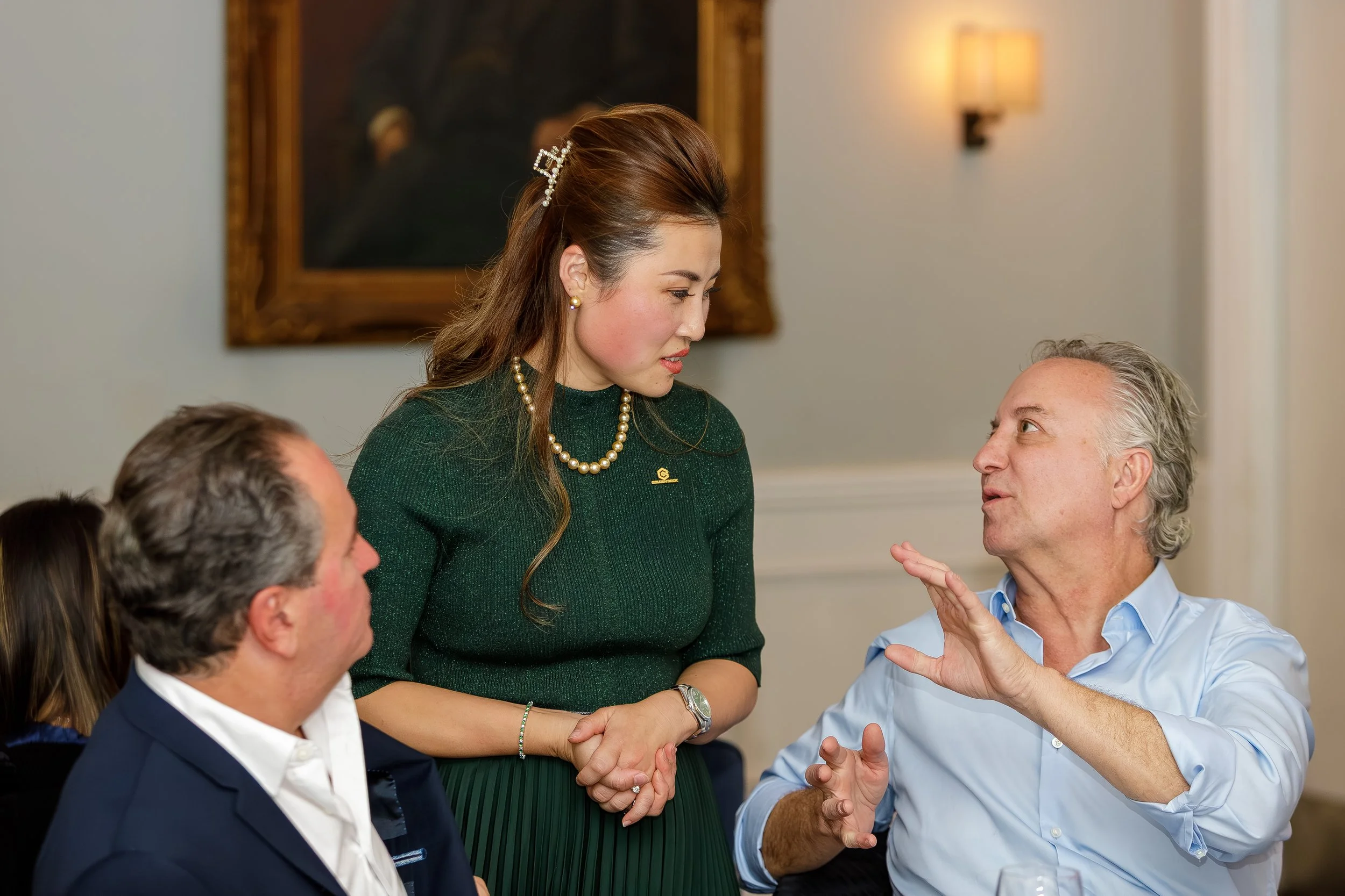 A woman in a green sweater and pearl necklace is standing while a man in a light blue shirt talks to her, seated at a round table during a formal gathering.