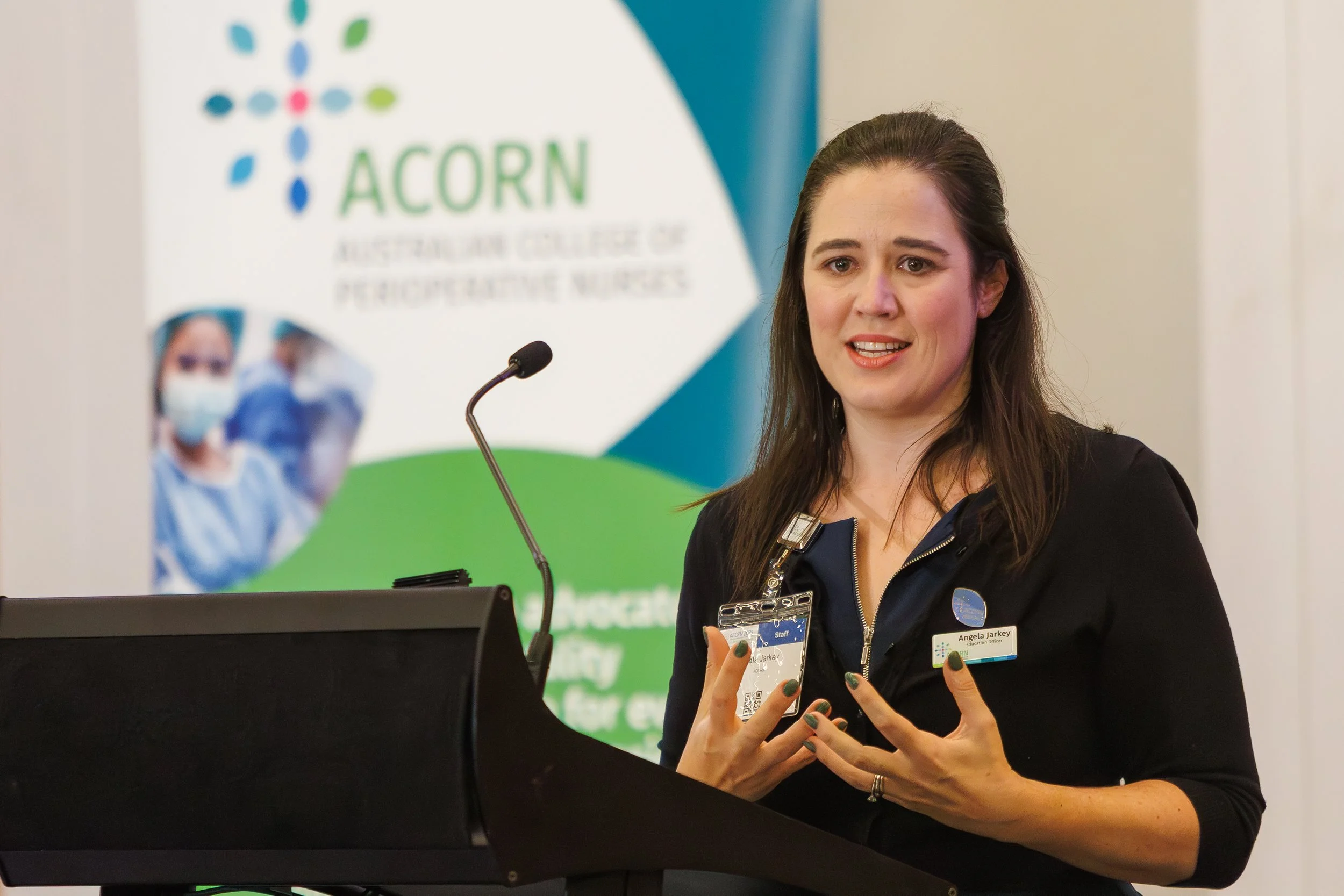 A woman with long brown hair wearing a black jacket and a badge stands at a podium, speaking with her hands gesturing. Behind her, there is a banner with the logo and name of ACORN, Australian College of Perioperative Nurses.