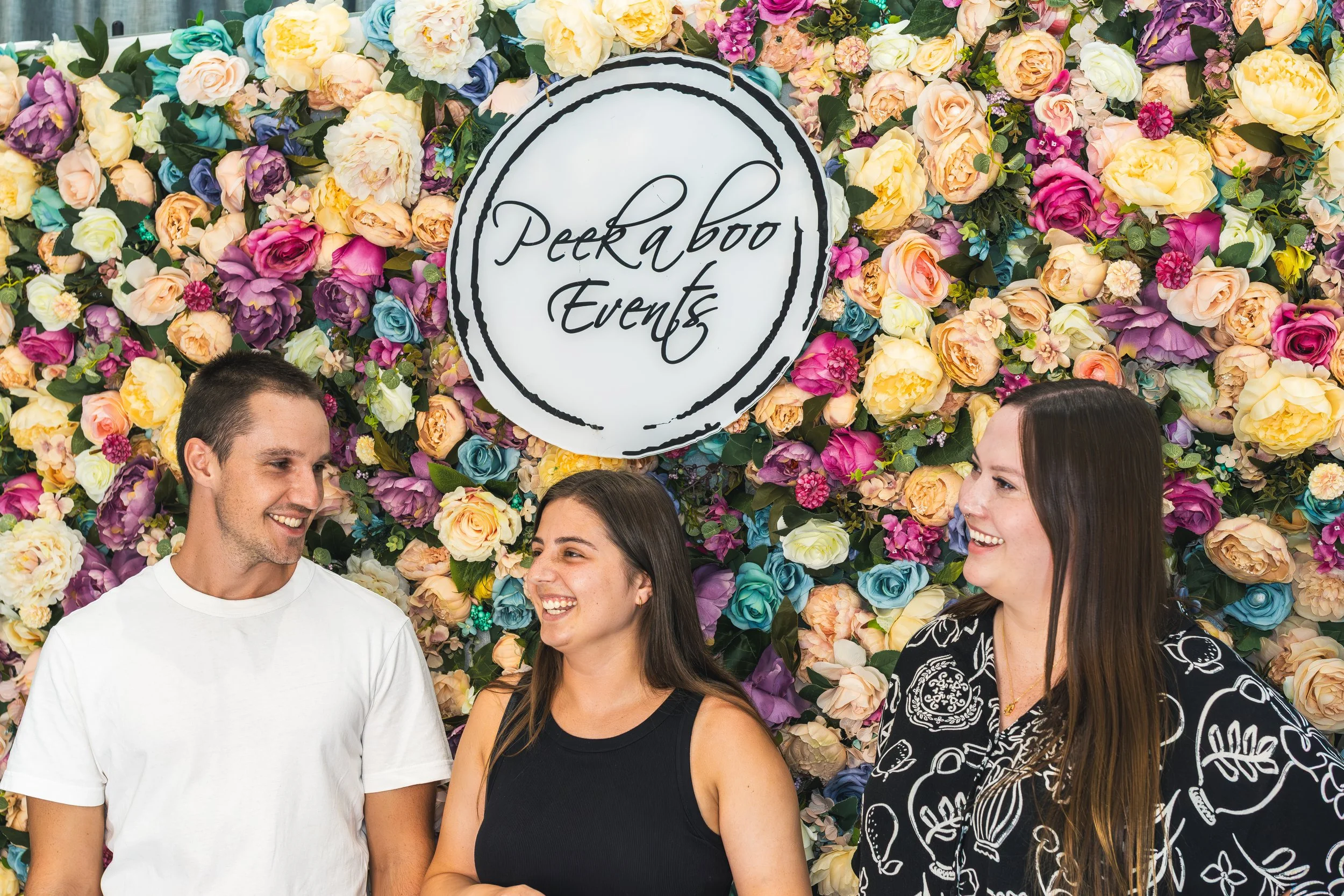 A group of four people smiling and talking in front of a colorful wall of flowers with a sign that says 'Peekaboo Events'.