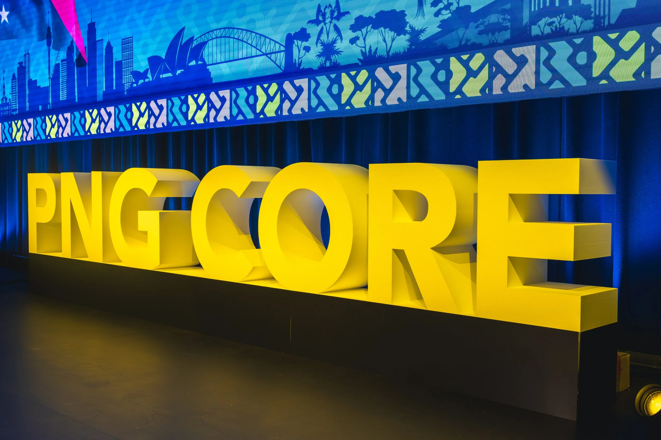 Large yellow 3D letters spell out 'NG CODE' on a black stage, with a vibrant blue and green cityscape and nature-themed artwork in the background.