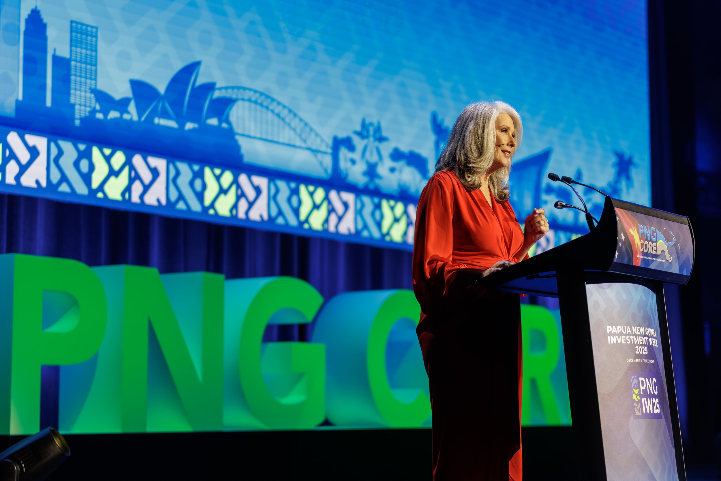 A woman with gray hair wearing a red dress speaking at a podium during a conference with a coral background. Behind her is a large screen displaying a blue image of skyline and landmark shapes, and large green 3D letters spelling 'DNC'.