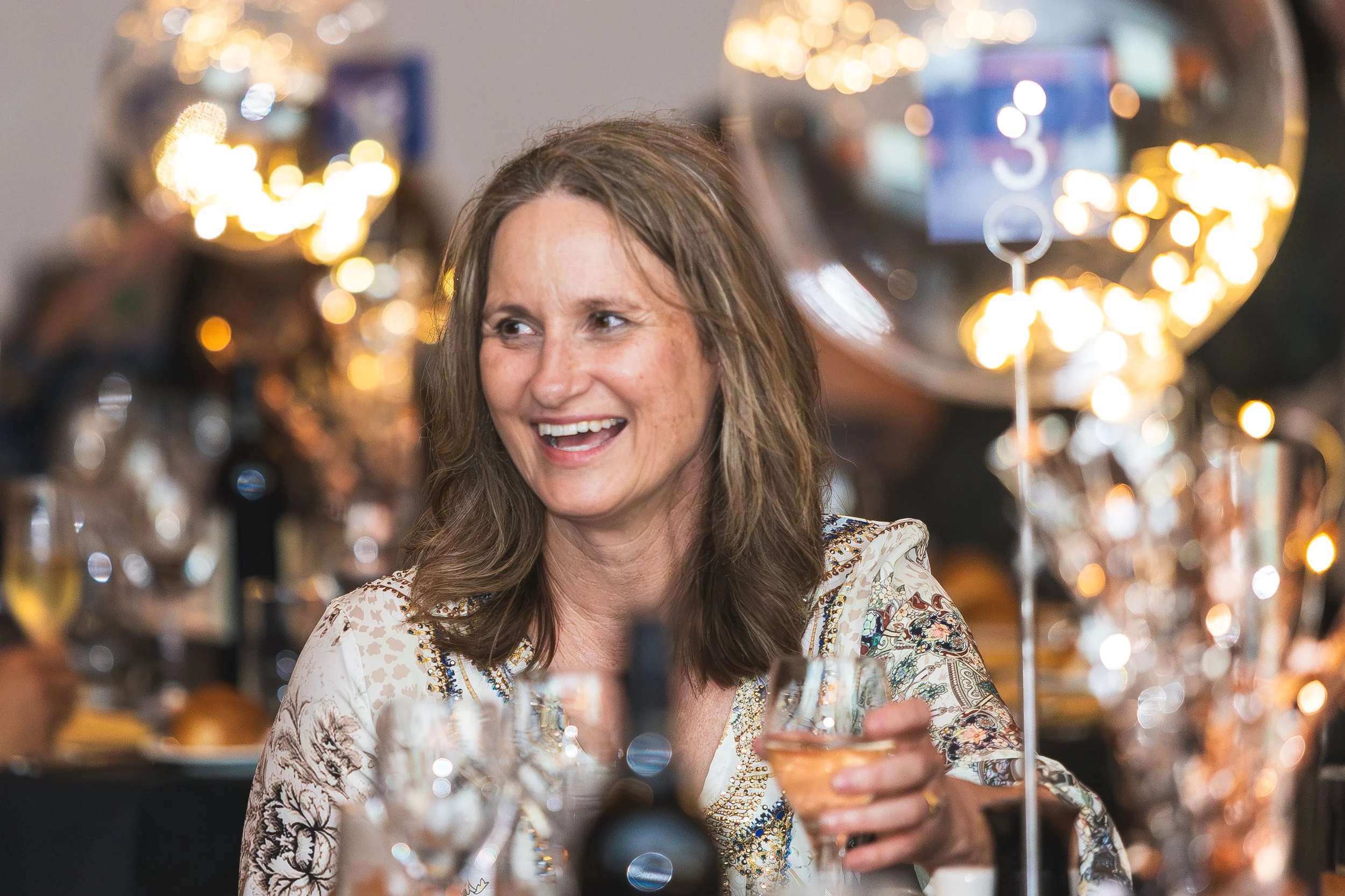 Woman with long brown hair smiling while holding a cocktail glass at a party or celebration, with blurred warm decorative lighting and reflections in the background.