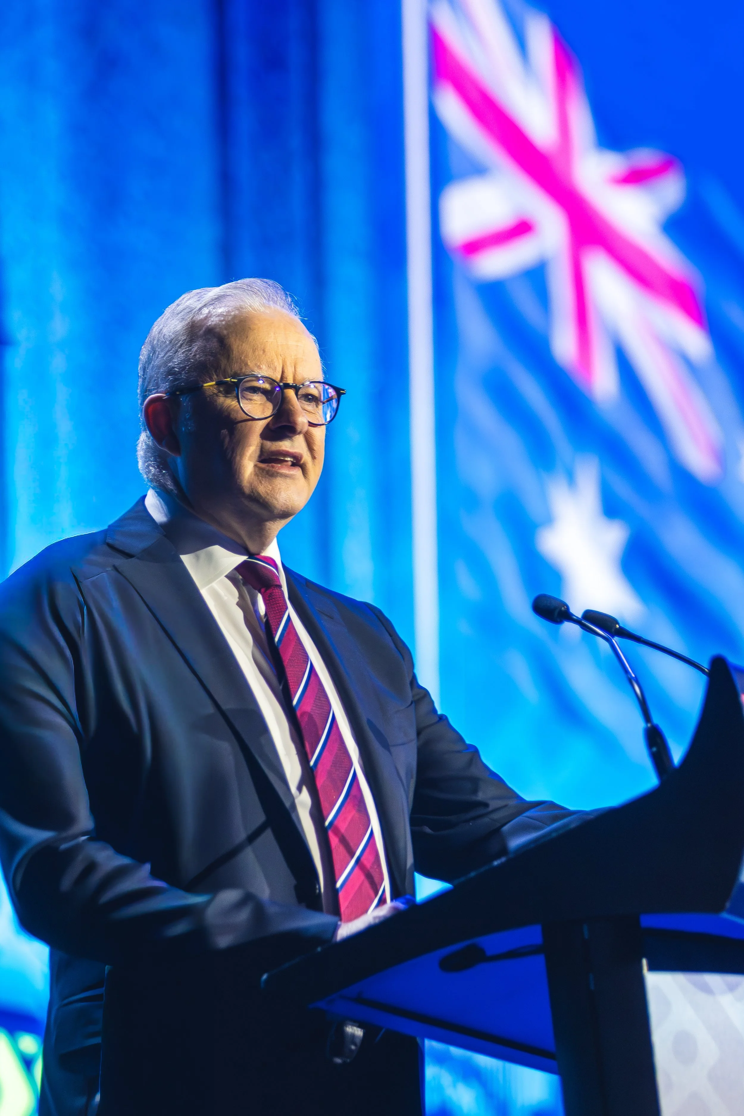 An elderly man with gray hair and glasses speaking at a podium with a microphone, in front of a blue background with a stylized Union Jack flag.