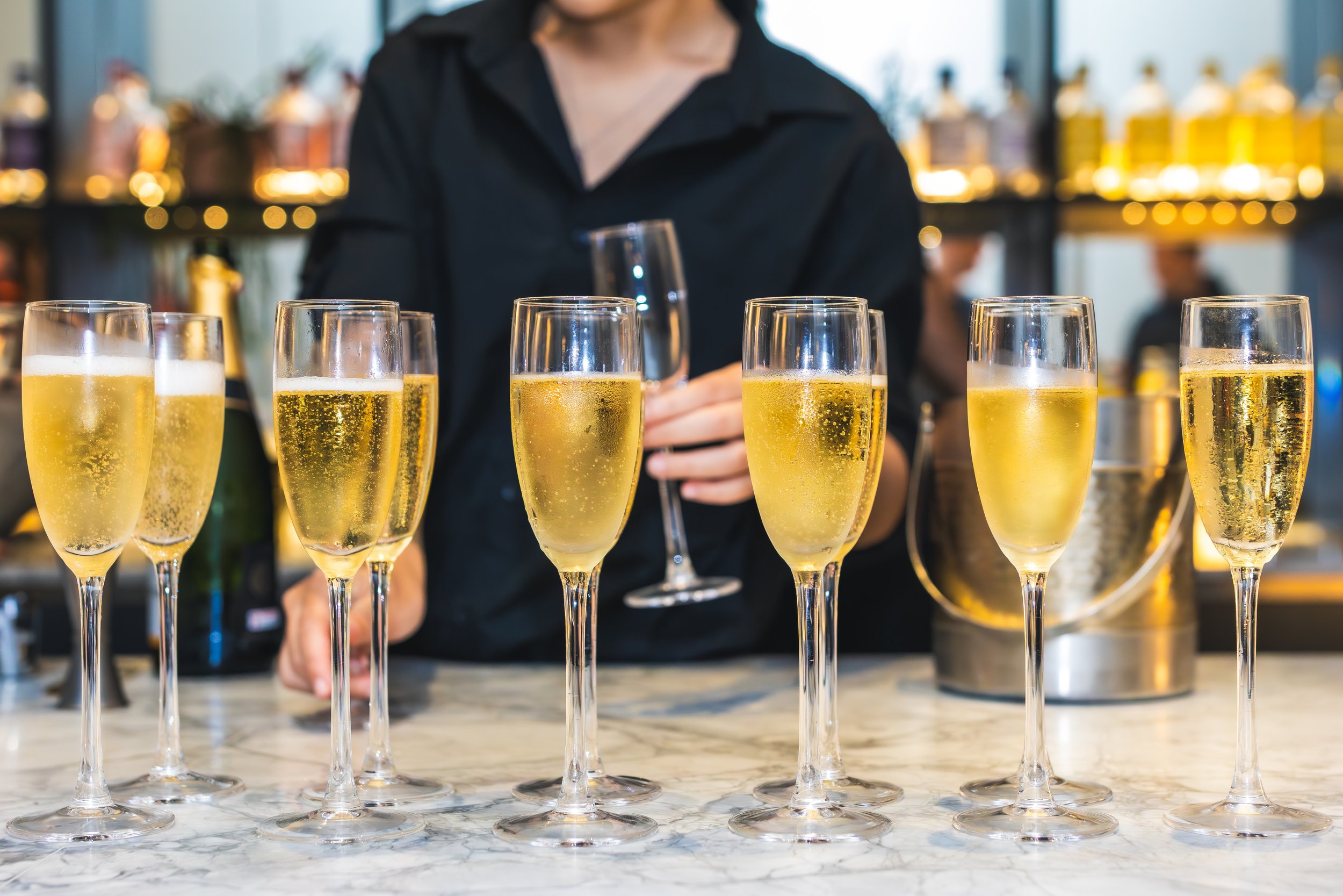 Multiple glasses of champagne on a bar counter with a bartender in the background serving a glass.