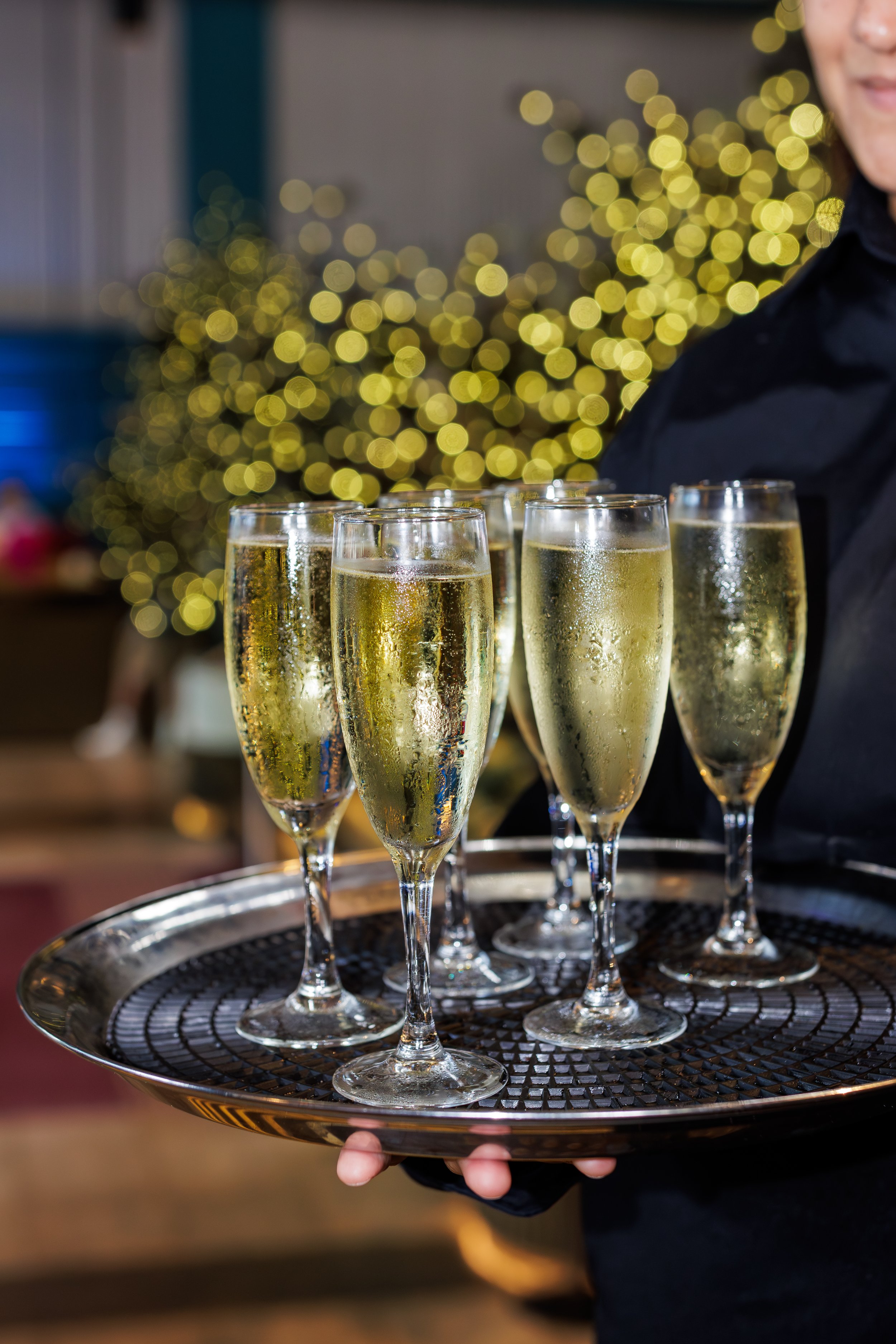 A tray of seven flutes of champagne with condensation, garnished with ice, held by a server in a black outfit. Christmas lights are blurred in the background.