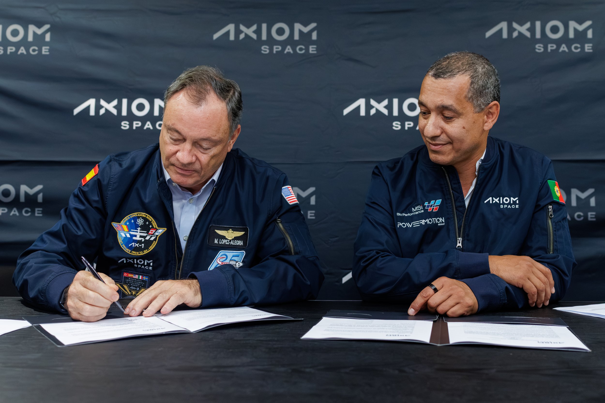 Two men in astronaut uniforms seated at a table, signing documents with an Axiom Space backdrop behind them.