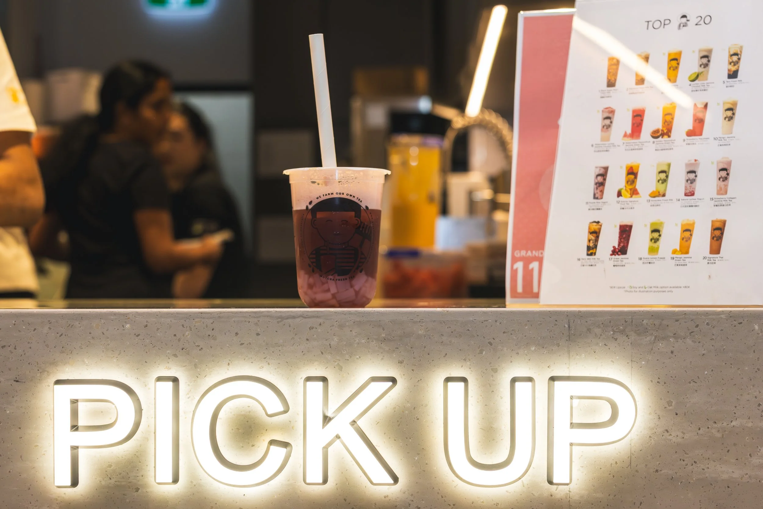 A cup of bubble tea on a counter with a menu in the background. The bubble tea is pink with tapioca pearls at the bottom, a white straw, and a logo featuring a smiling cartoon girl.