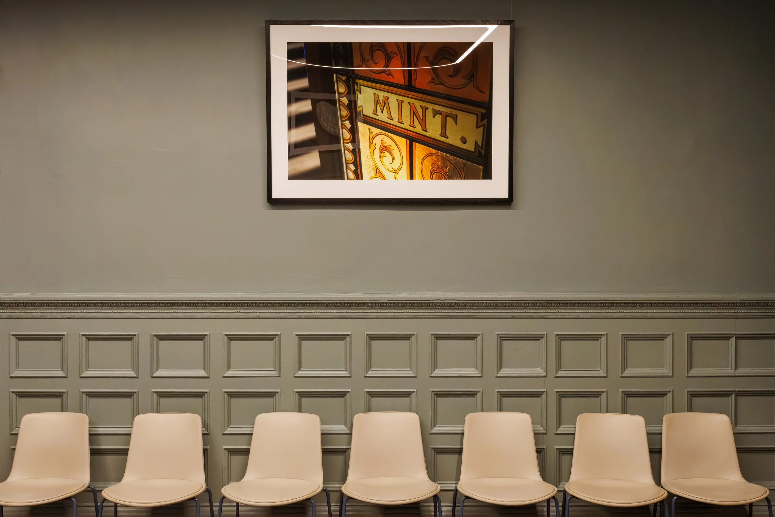 Empty beige chairs in front of a gray wainscoted wall with a framed vintage-style sign reading 'MINT' and a decorative trim near the ceiling.
