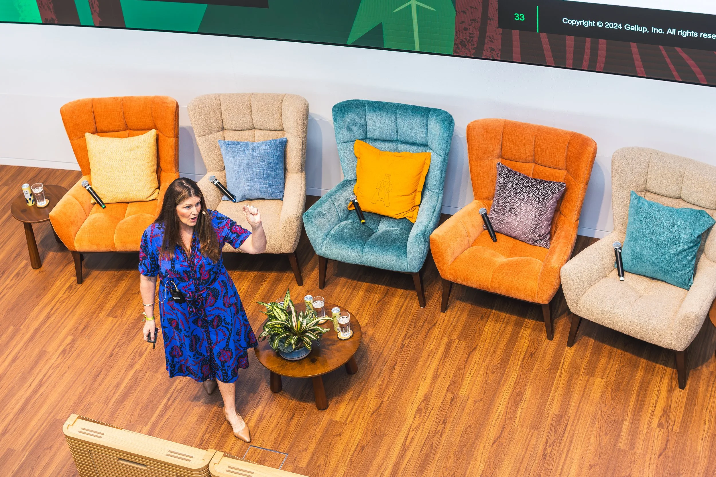 A woman with long brown hair wearing a blue patterned dress is standing in front of a row of colorful high-backed chairs, holding a microphone in one hand and gesturing with her other hand. The chairs are orange, beige, teal, orange, and beige, each 