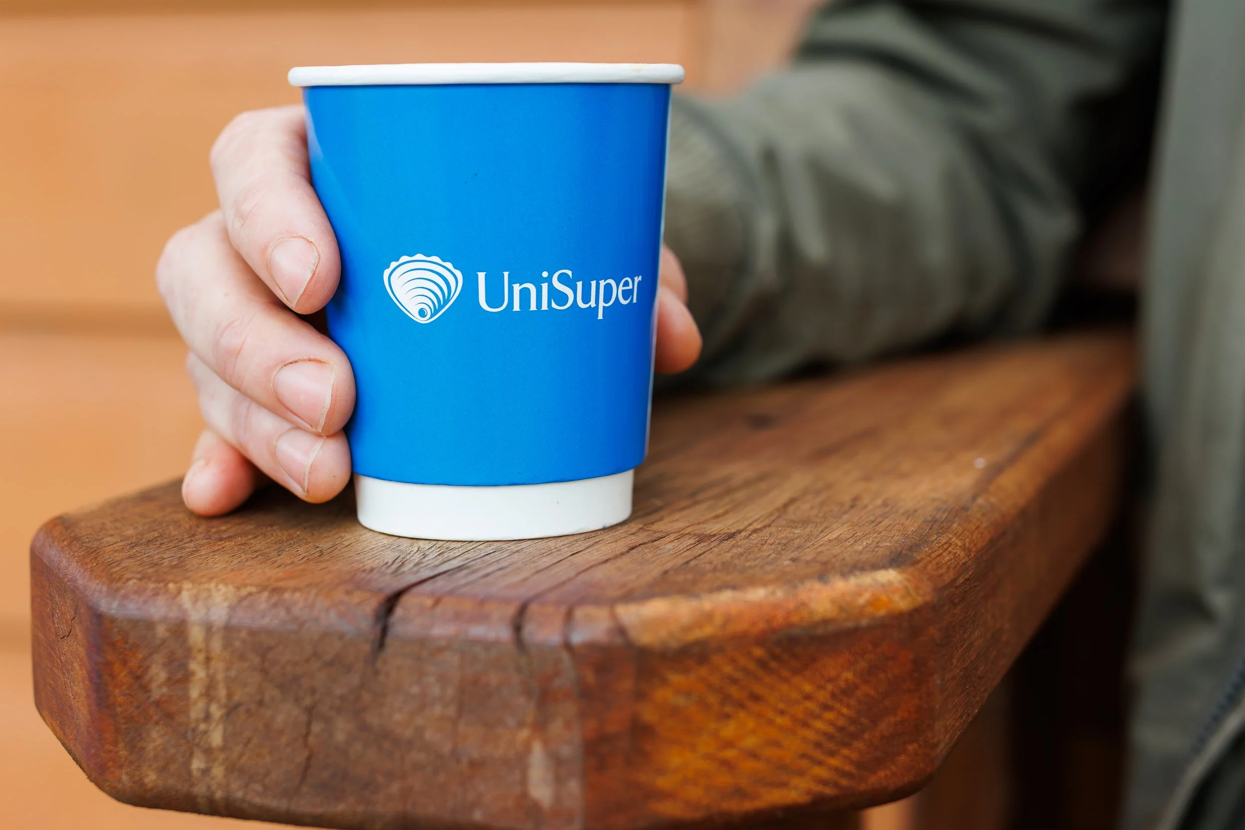 A hand holding a blue paper cup with the UniSuper logo, resting on a wooden surface.