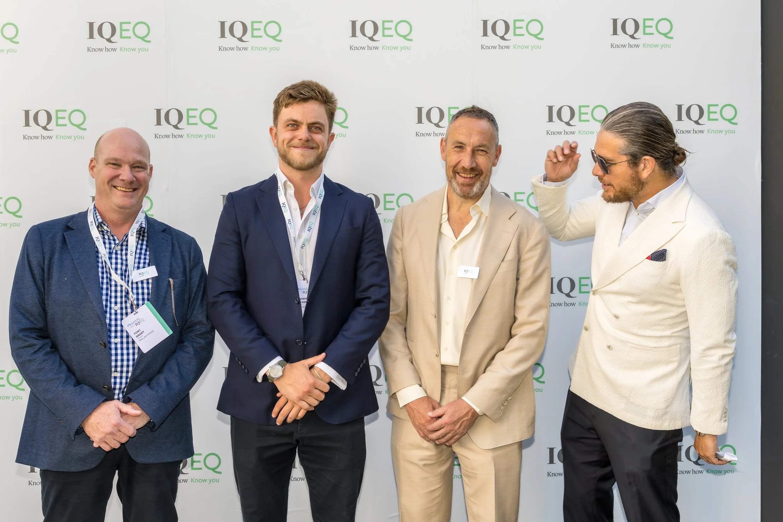 Four men standing in front of an IQEQ branded backdrop, dressed in business attire, smiling and engaging in conversation.