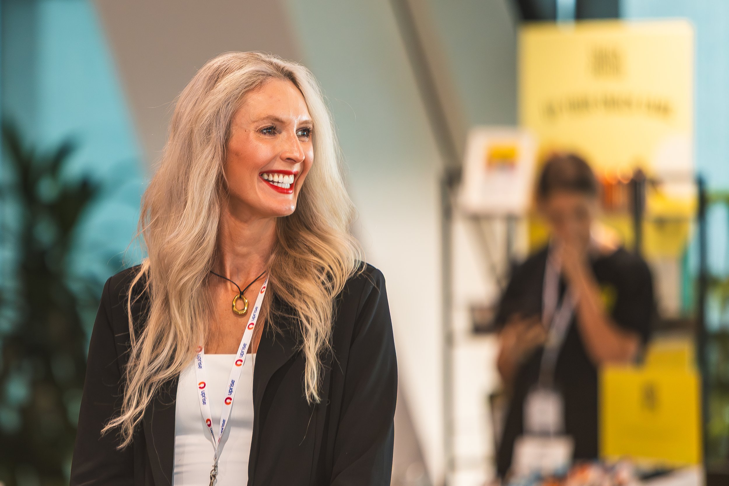 A woman with long blonde hair smiling, wearing a black blazer and white top, walking in an indoor setting with colorful signs and other people in the background.
