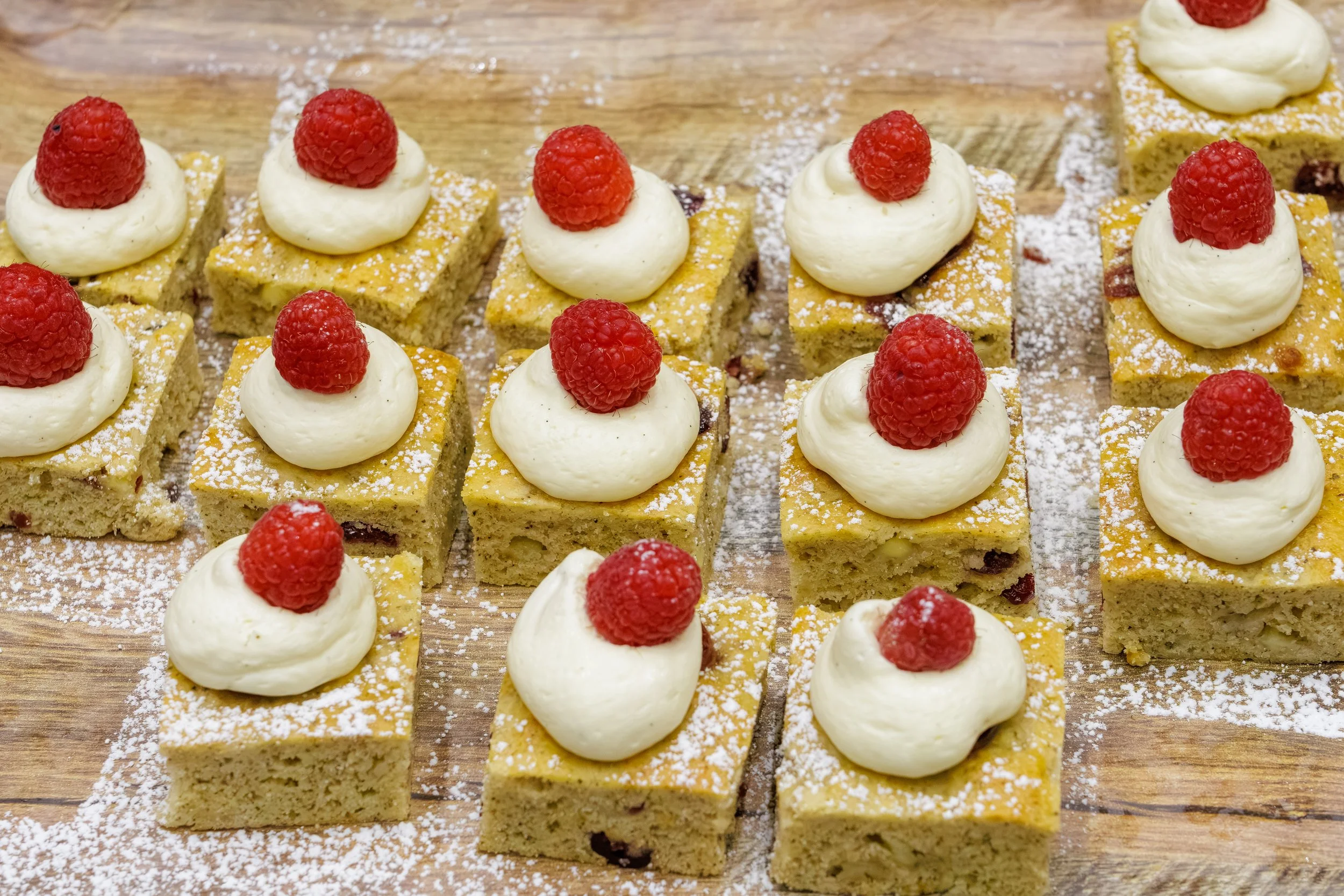 Squares of cake topped with whipped cream and a raspberry, dusted with powdered sugar, arranged on a wooden surface.
