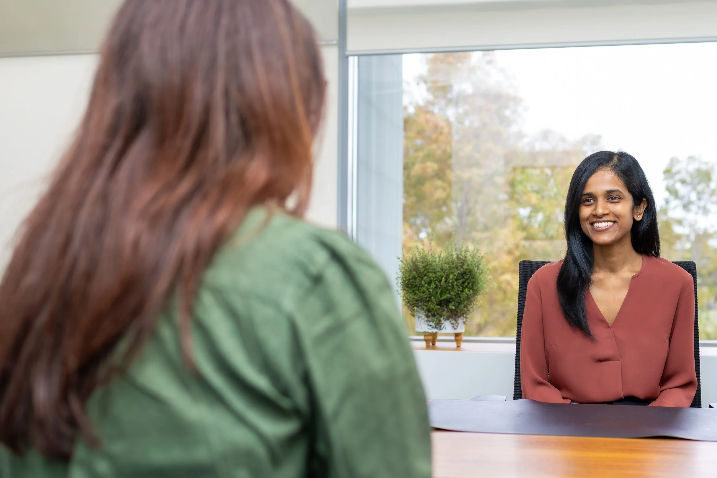 Two women are in a meeting room; one woman with black hair and a reddish-brown blouse is smiling at the other woman with red hair and a green jacket. They are seated across from each other at a table, with a window showing trees outside in the backgr